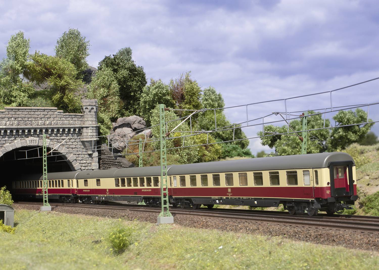 Modelový vlak v červeno-béžové barvě vyjíždí z tunelu na zelené, zalesněné trati pod oblohou pokrytou mraky.