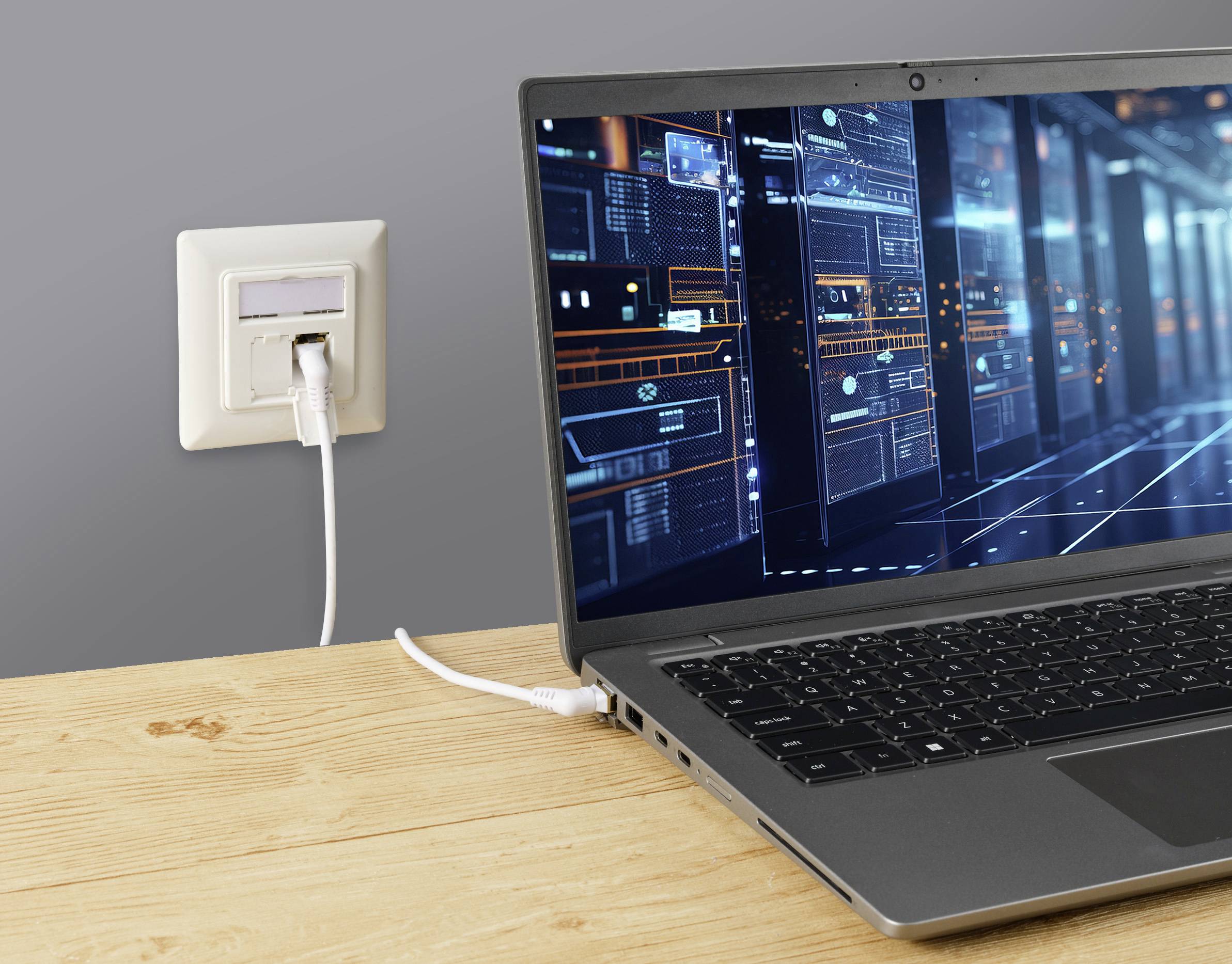 Laptop na stole je připojen kabelem k síťové zásuvce ve zdi. Na obrazovce jsou viditelné digitální síťové diagramy.