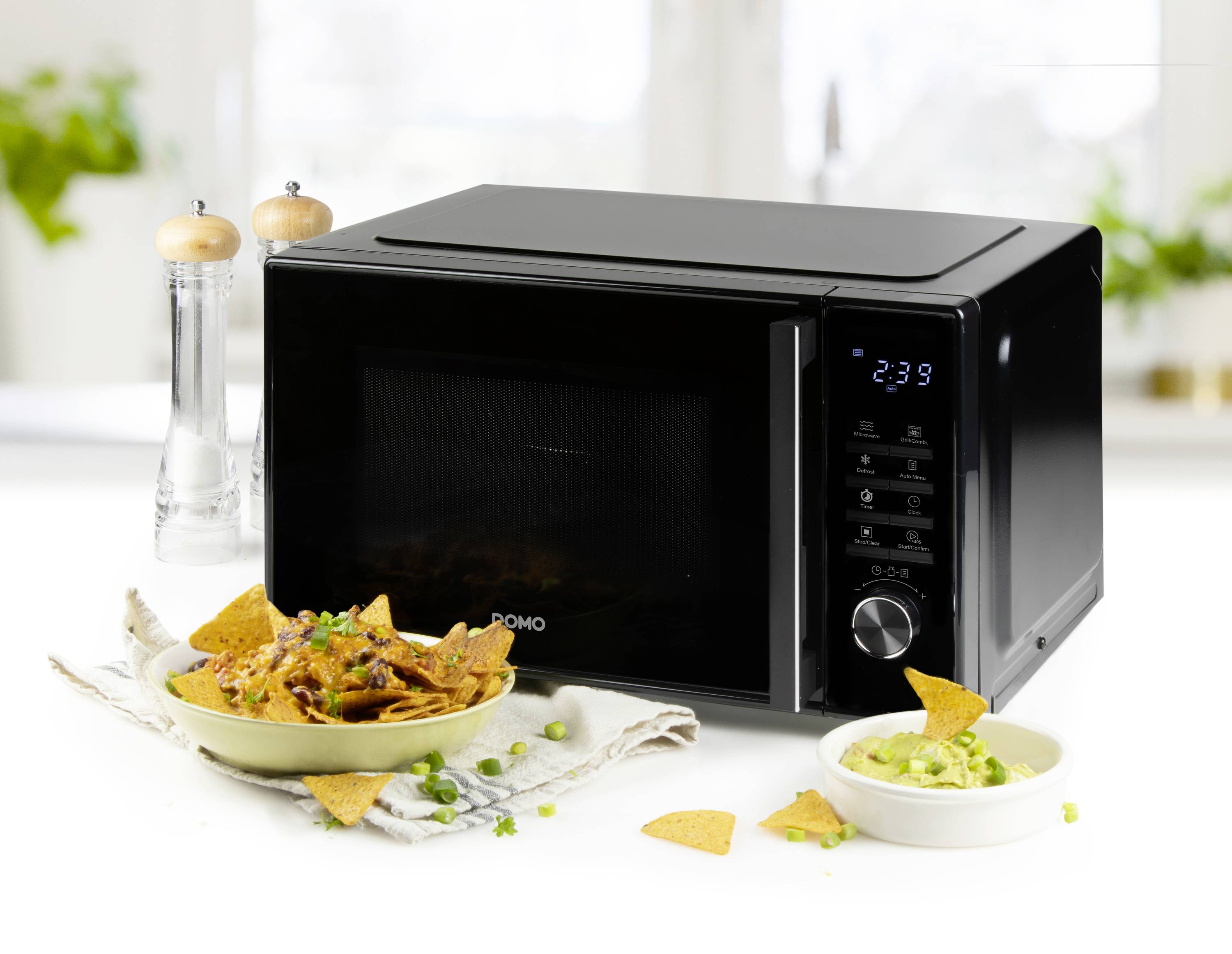 Černá mikrovlnka na bílém stole, obklopená slánkou, pepřenkou a talířem s nachos a guacamole.