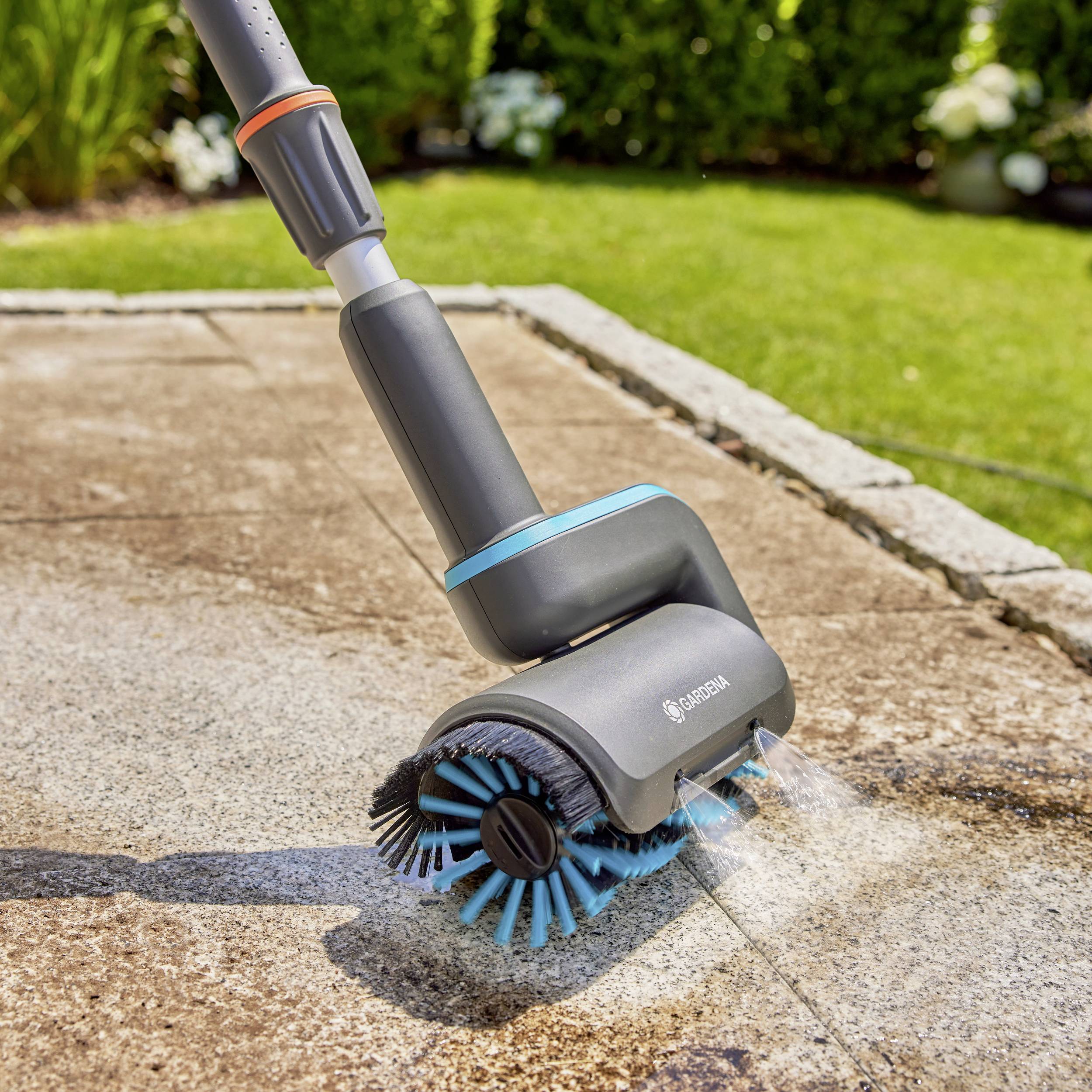 Elektricky poháněný čistič teras čistí chodník v zahradě pokrytý kamennými dlaždicemi. V pozadí jsou zelené keře.