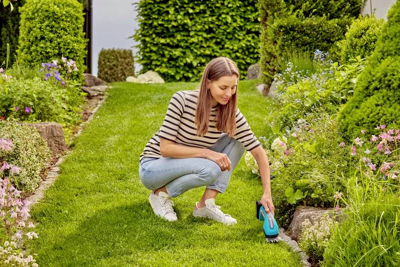 Žena dřepí na zahradní cestičce a mezi květinami a keři používá senzor vlhkosti půdy, aby zkontrolovala úroveň hydratace rostlin.