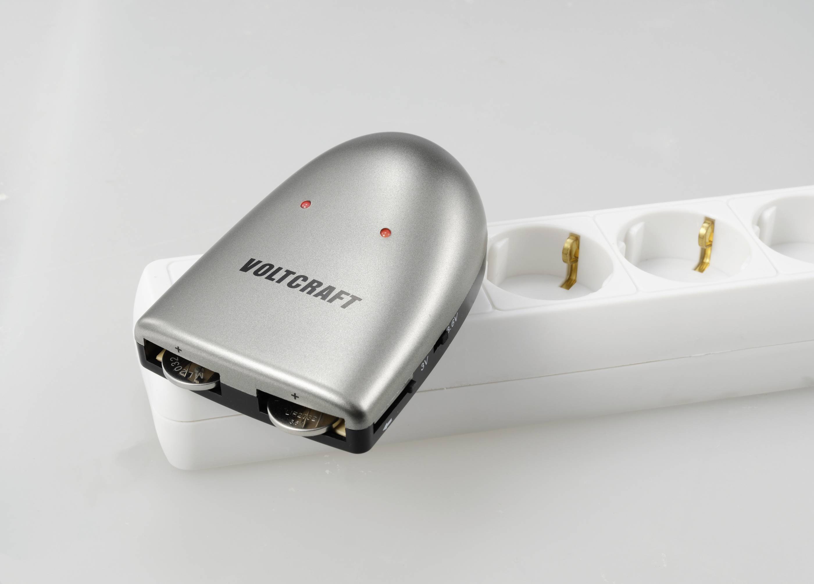 Na bílém rozvodném elektrickém panelu leží stříbrný měřič proudu značky Voltcraft.