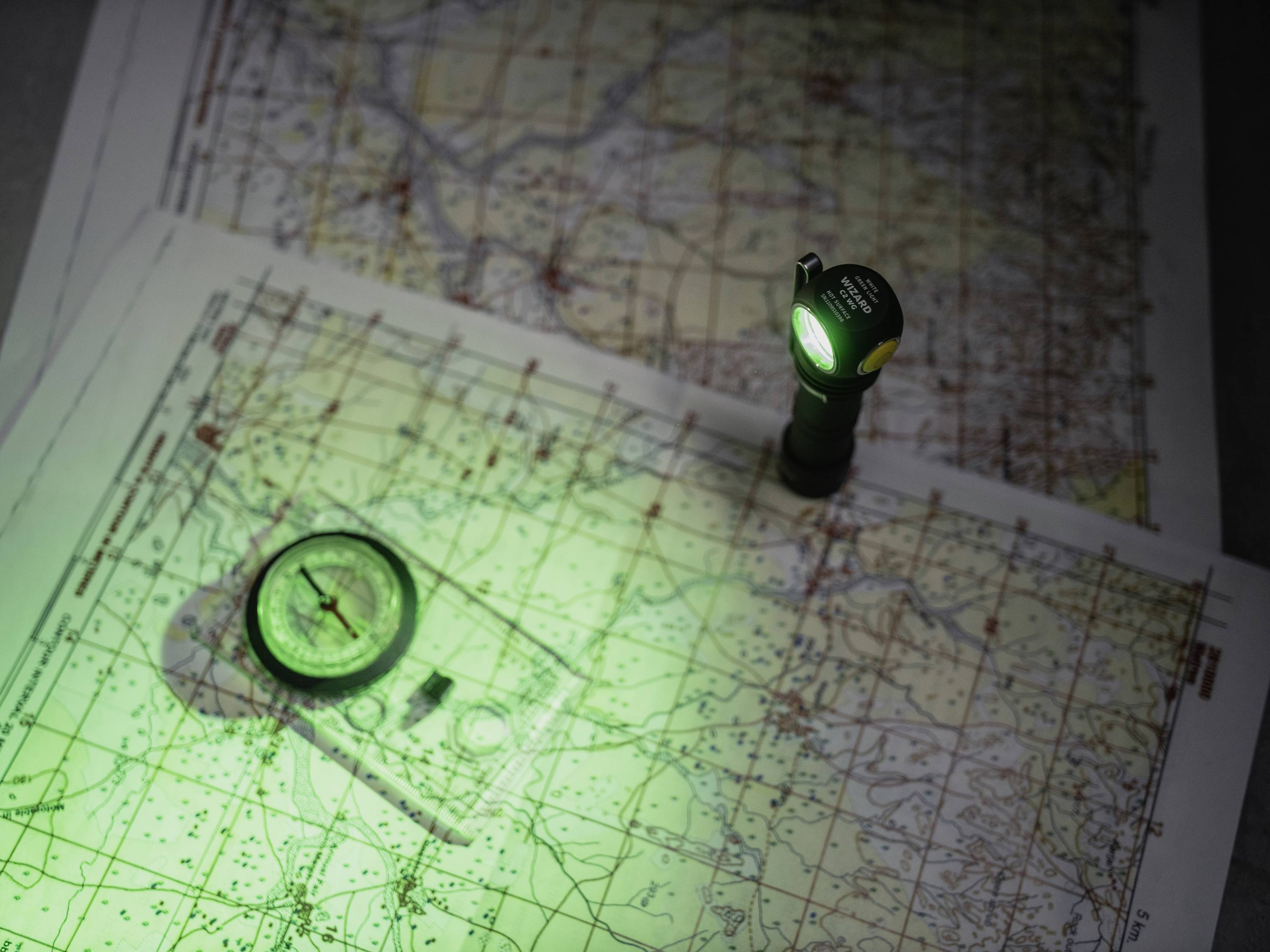 Mapy s kompasem a baterkou. Zelené světlo baterky osvětluje mapy s topografickými detaily a trasami.