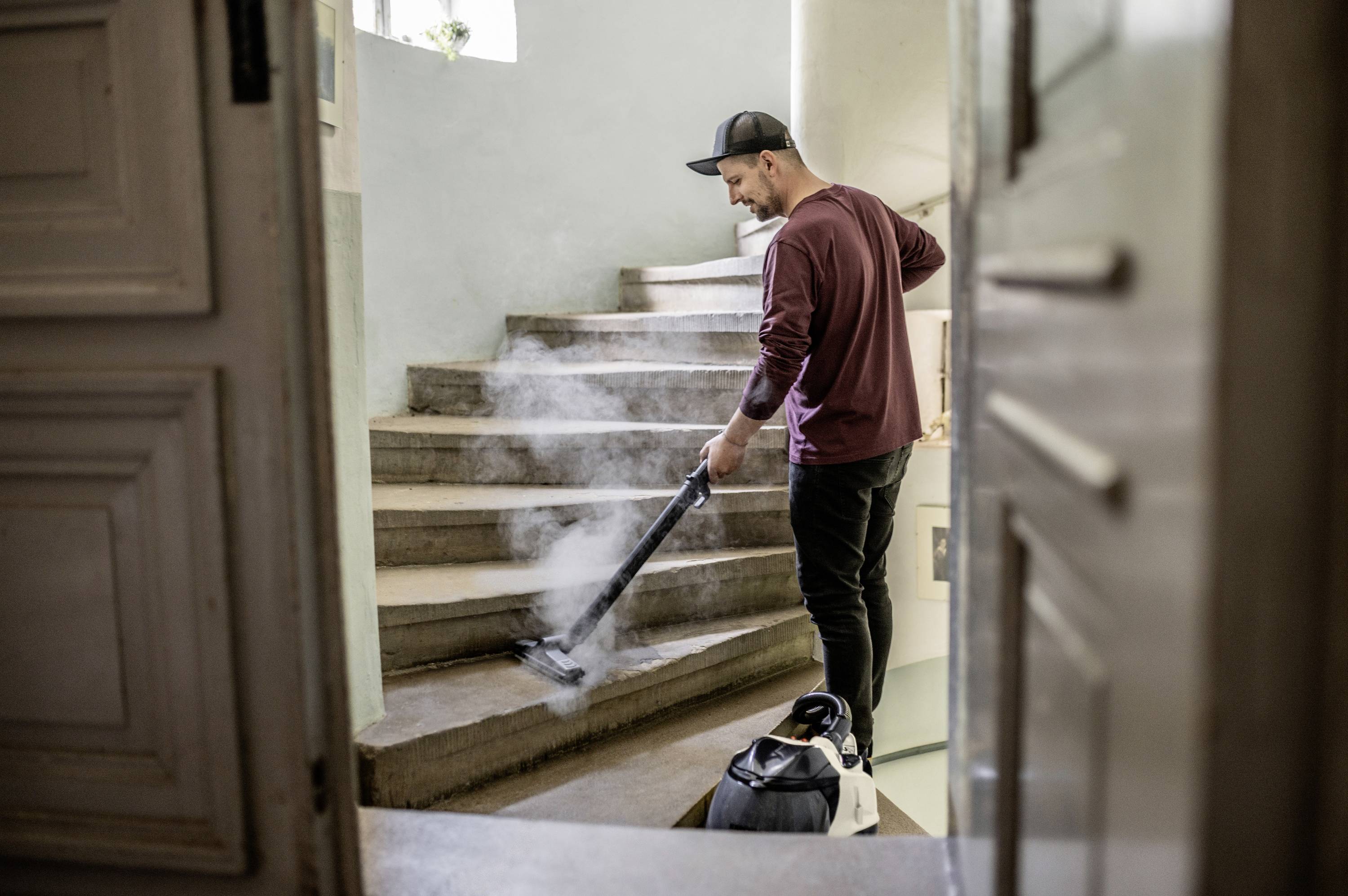 Osoba čistí schody parním vysavačem. Má na sobě volnočasové oblečení a baseballovou čepici. Prostředí působí domácky.