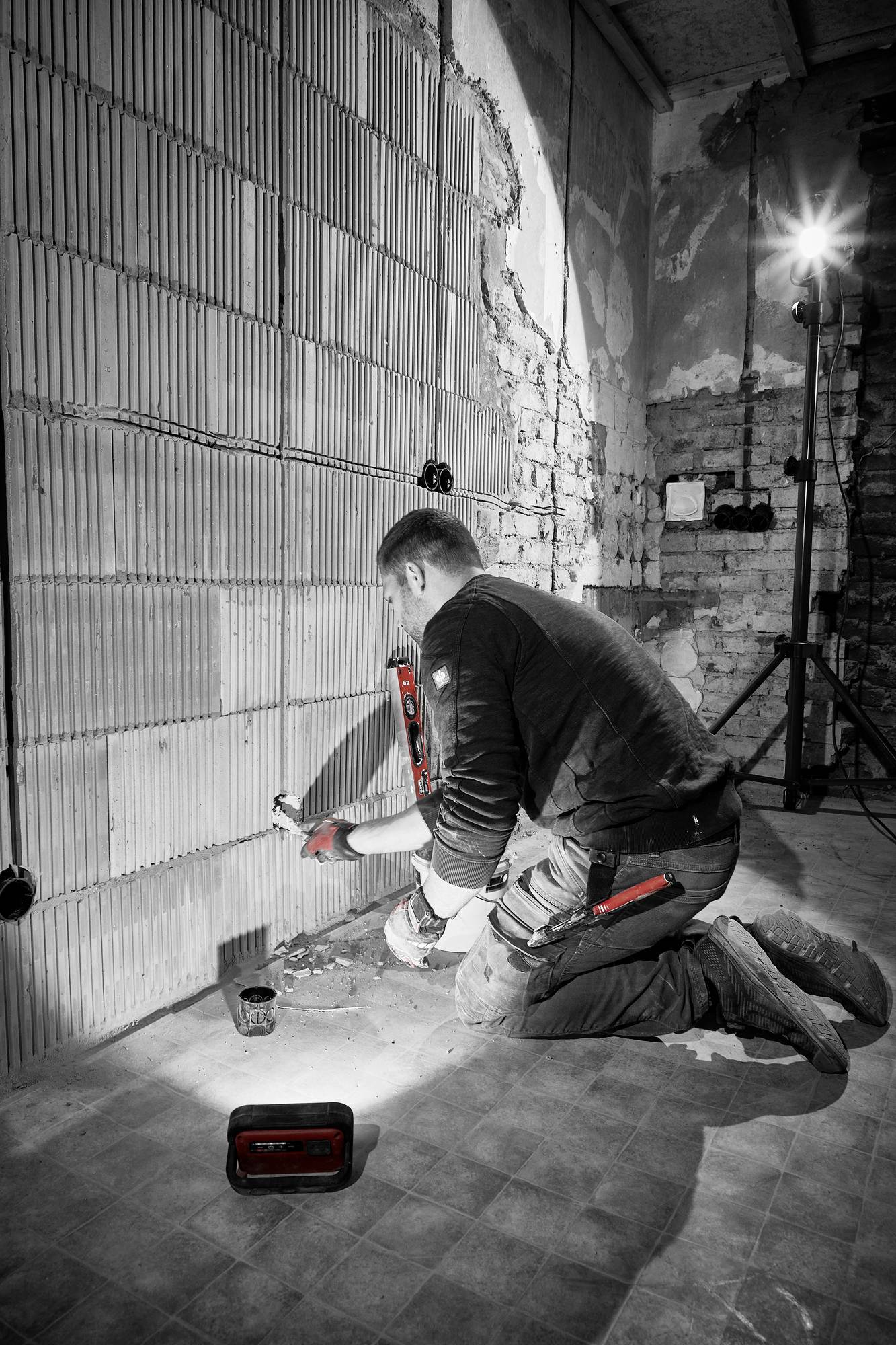 Dělník klečí před stěnou v nedokončené stavbě a pracuje s nástroji na elektroinstalaci. Stavební reflektor osvětluje prostor.