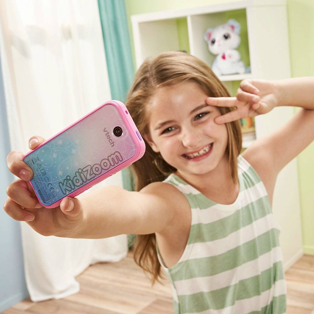 Dívka se usmívá do kamery a ukazuje znamení vítězství, zatímco si s růžovým dětským fotoaparátem pořizuje selfie.