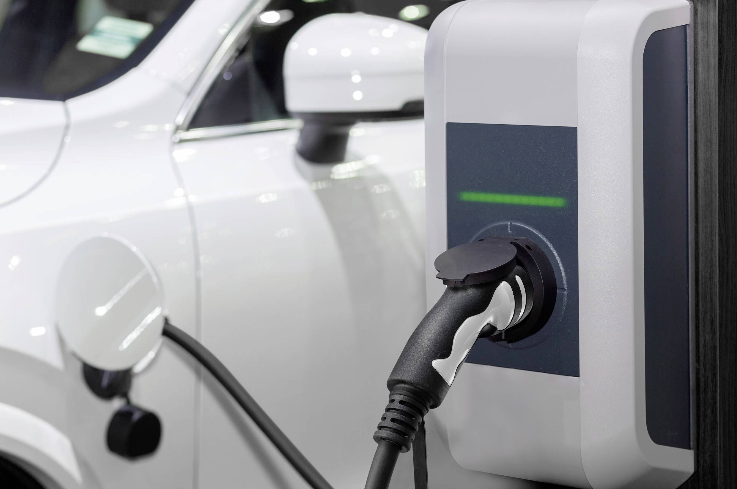 Bílý elektromobil se nabíjí na nabíjecí stanici. V popředí je zřetelně viditelný nabíjecí kabel.