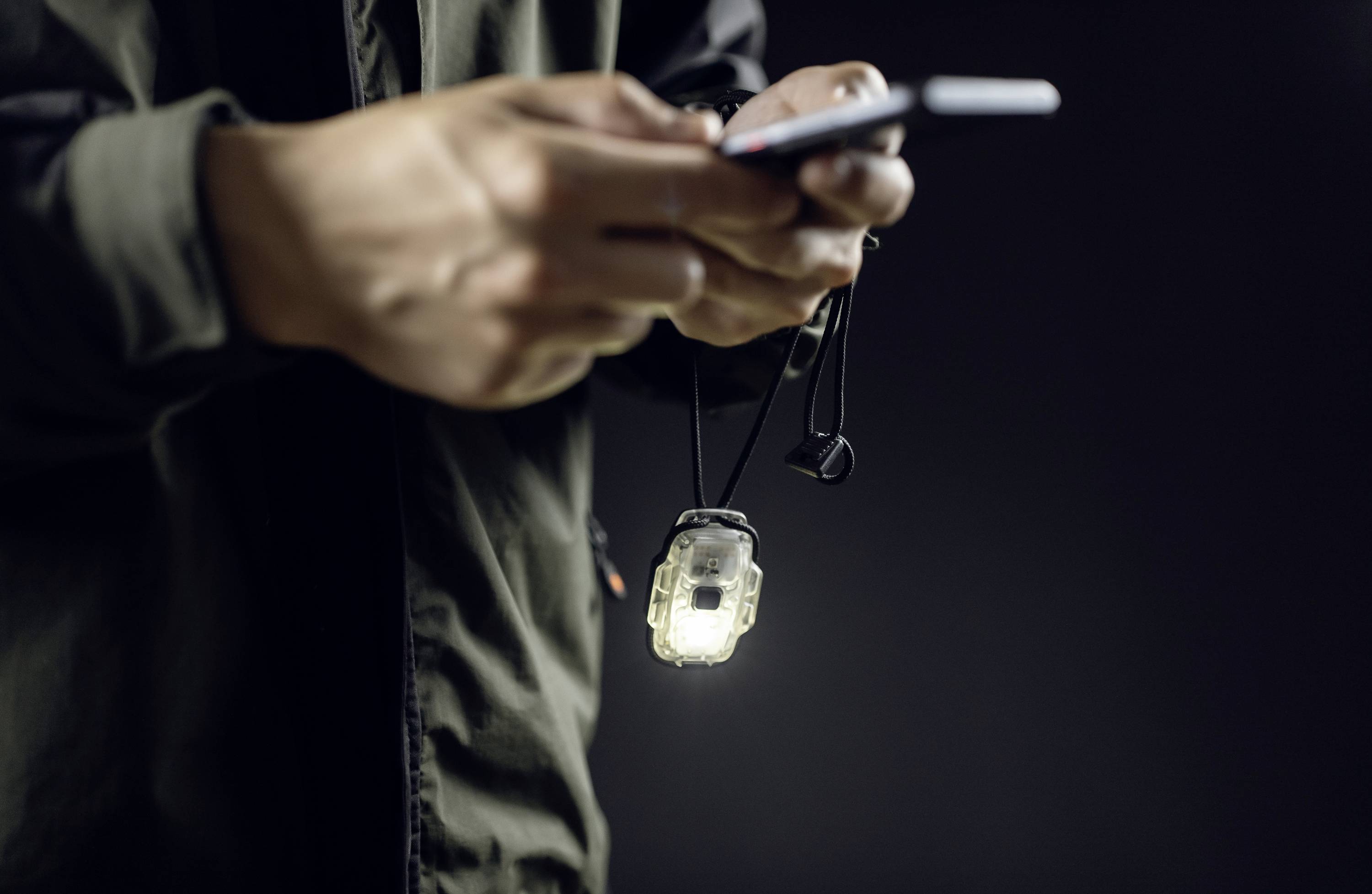 Detail záběru rukou svírající smartphone. Na šňůrce visí svítící baterka. Pozadí je tmavé.