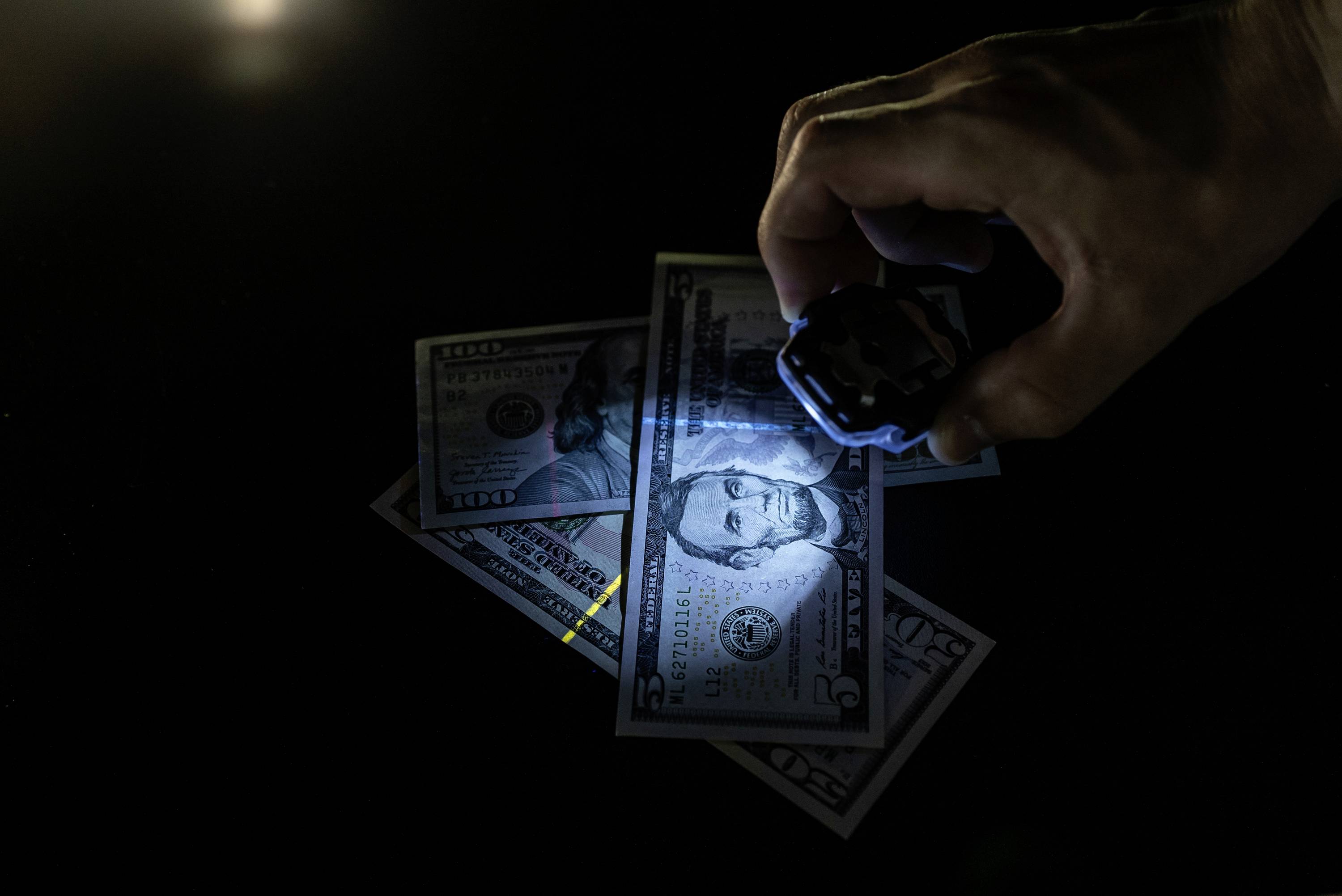 Ruka drží UV lampu nad několika bankovkami amerických dolarů, které zobrazují vyznačené bezpečnostní prvky.