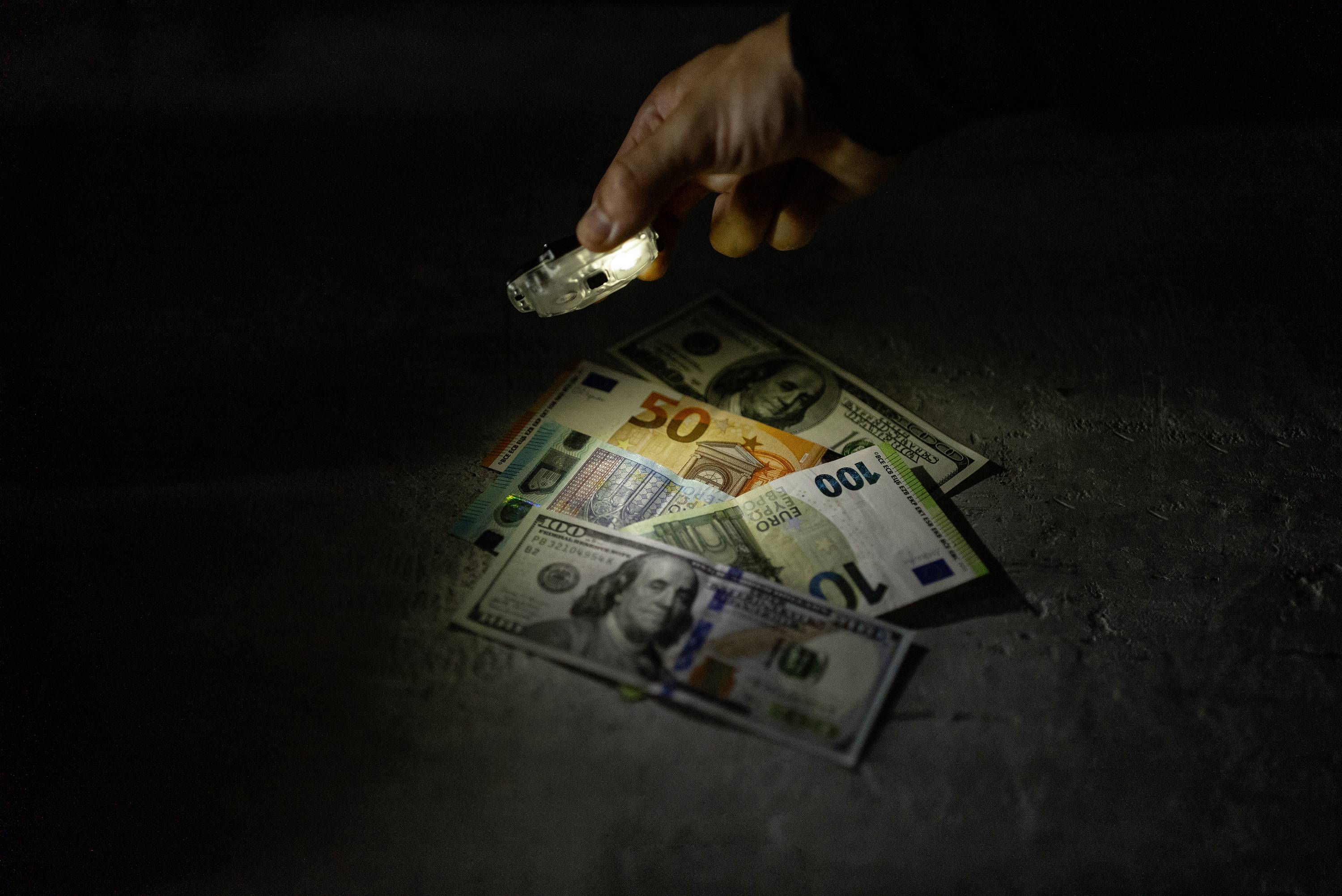 Ruka s baterkou osvětluje několik bankovek, včetně eur a amerických dolarů, na tmavém povrchu.