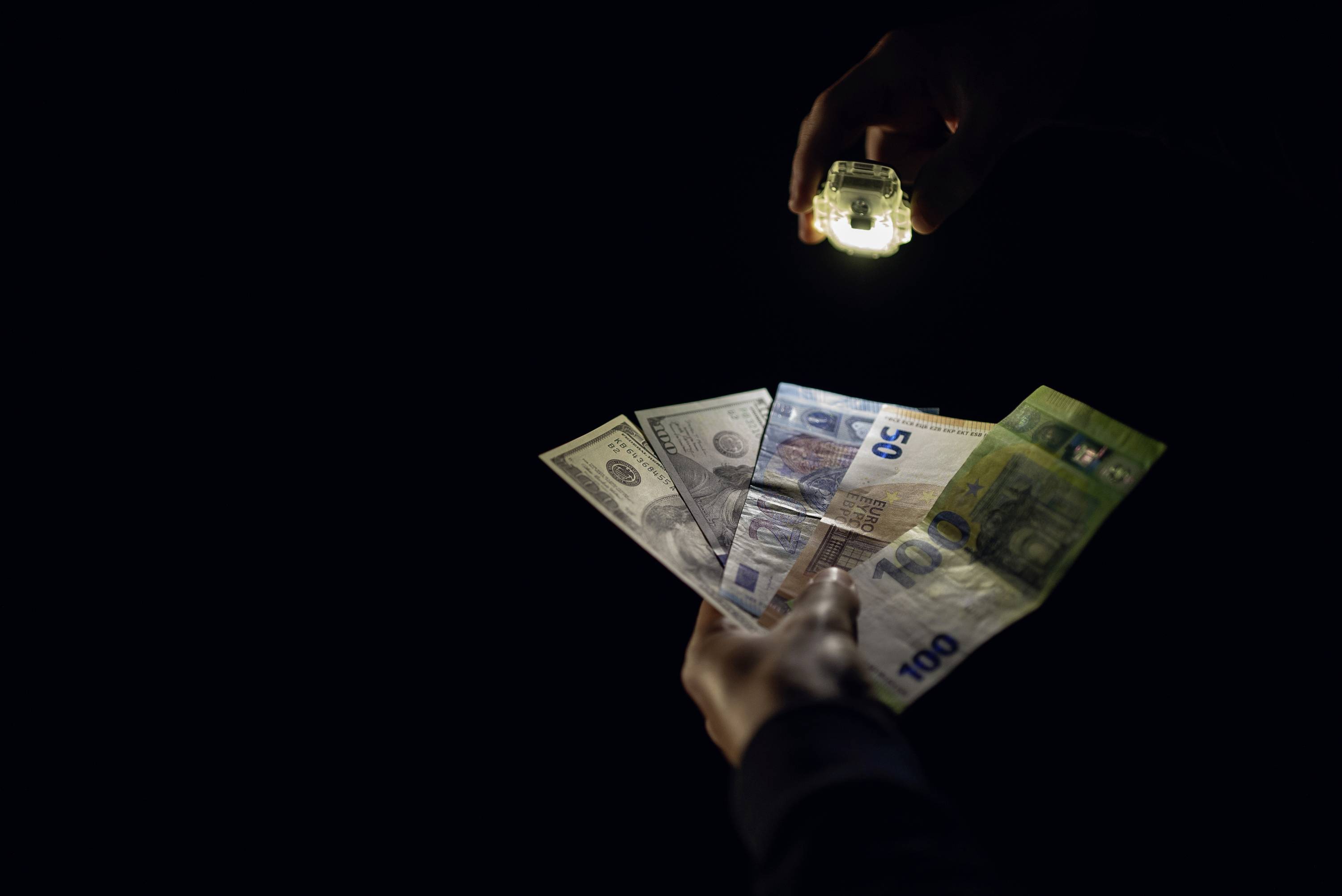 Jedna ruka drží různé bankovky, včetně amerických dolarů a eur, zatímco druhá ruka je osvětluje baterkou.