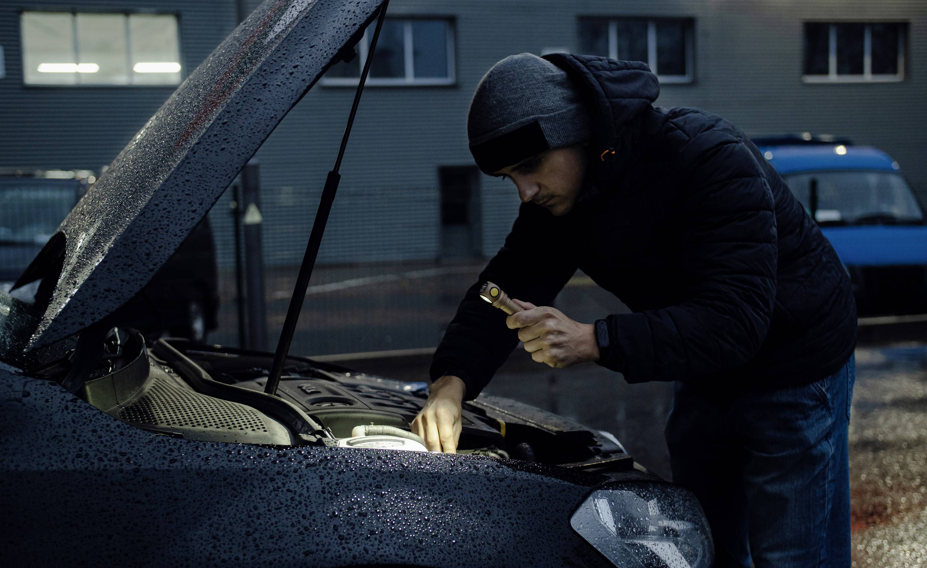Muž s kapucí prozkoumává motor zaparkovaného vozu baterkou za deště.