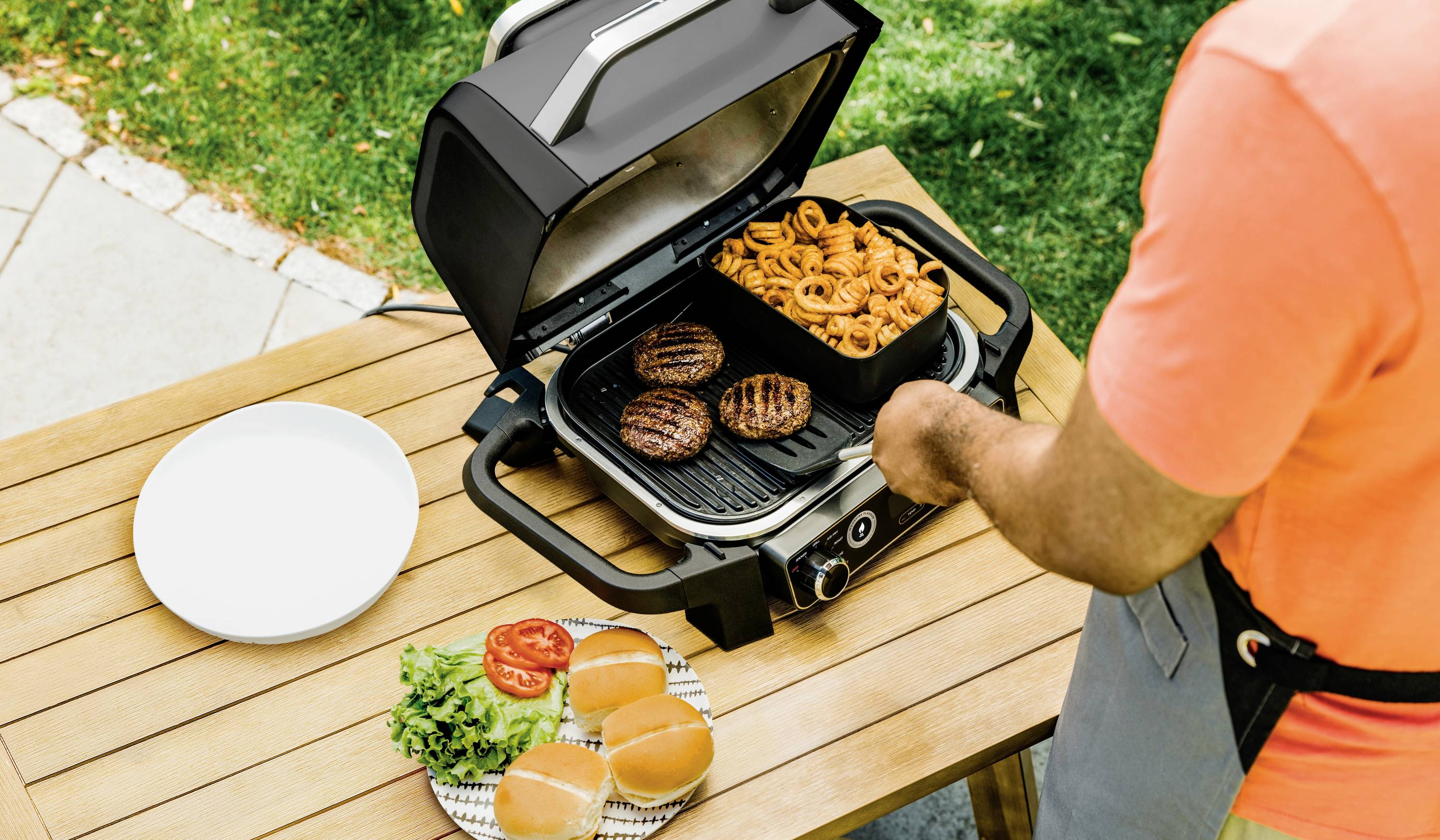 Muž griluje burgery a hranolky na plynovém grilu venku. Na stole leží hamburgerové bulky, salát, rajčata a prázdný talíř.