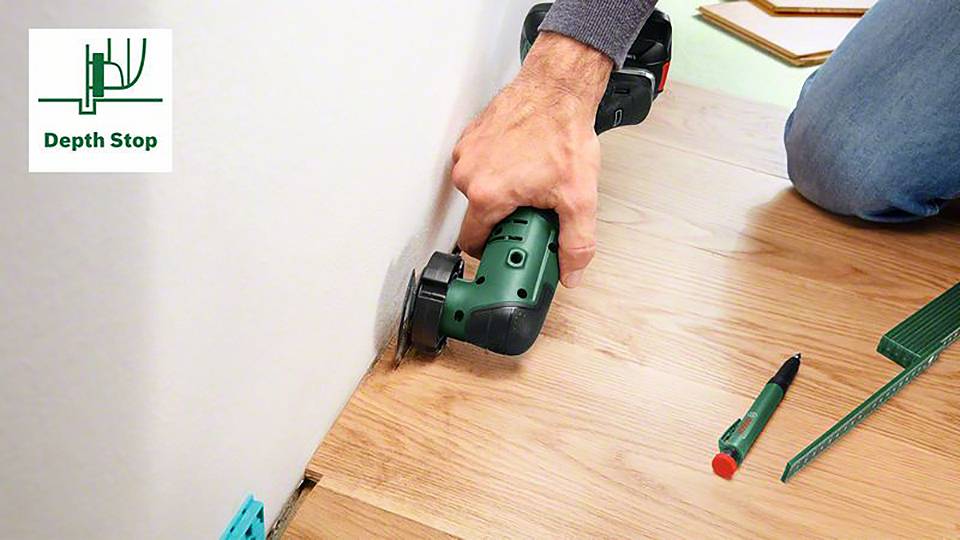 Jedna osoba používá elektrický nástroj k řezání dřevěné podlahy poblíž zdi. Na podlaze leží pero a pravítko.