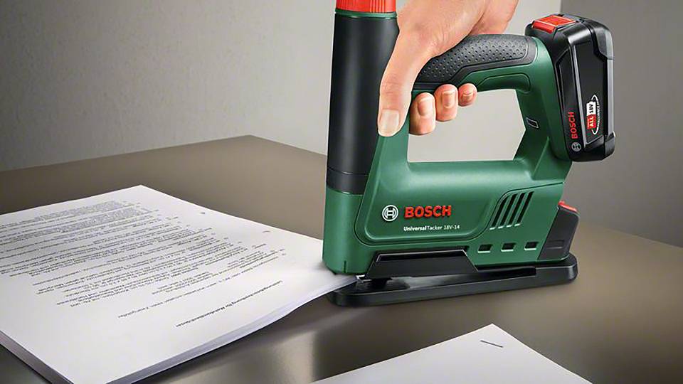 Jedna ruka drží zelený elektrický sponkovač Bosch, který sešívá papíry na stole.