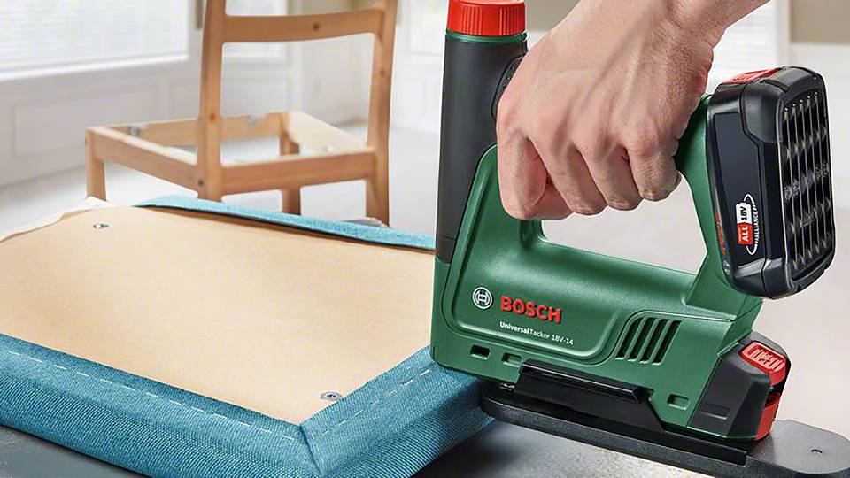 Osoba drží zelený Bosch sponkovač a upevňuje modrý textil na dřevěný rám. V pozadí je vidět židle.