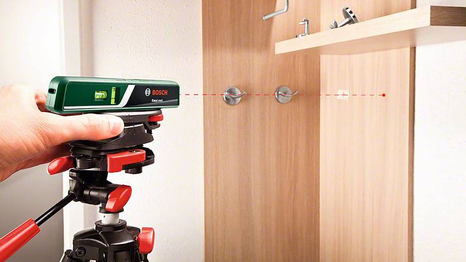 Jedna ruka drží laserové zařízení Bosch na stativu, které promítá červený laserový paprsek na dřevěnou zeď za účelem kontroly zarovnání.