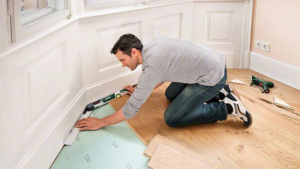 Muž klečí na dřevěné podlaze a pokládá izolační podložku podél stěny. Nástroje a materiály jsou připraveny.