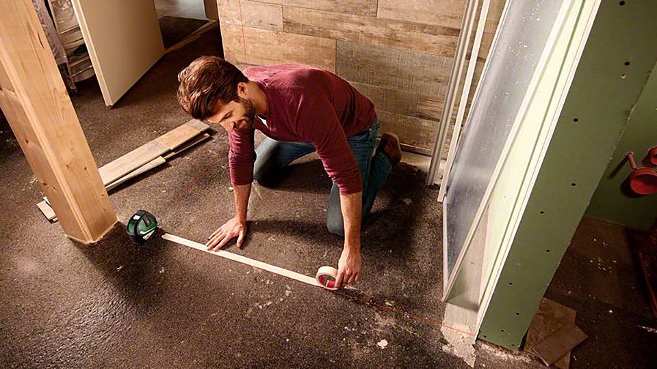 Jedna osoba klečí na podlaze a používá svinovací metr k měření vzdálenosti mezi dvěma stěnami při stavebním projektu.