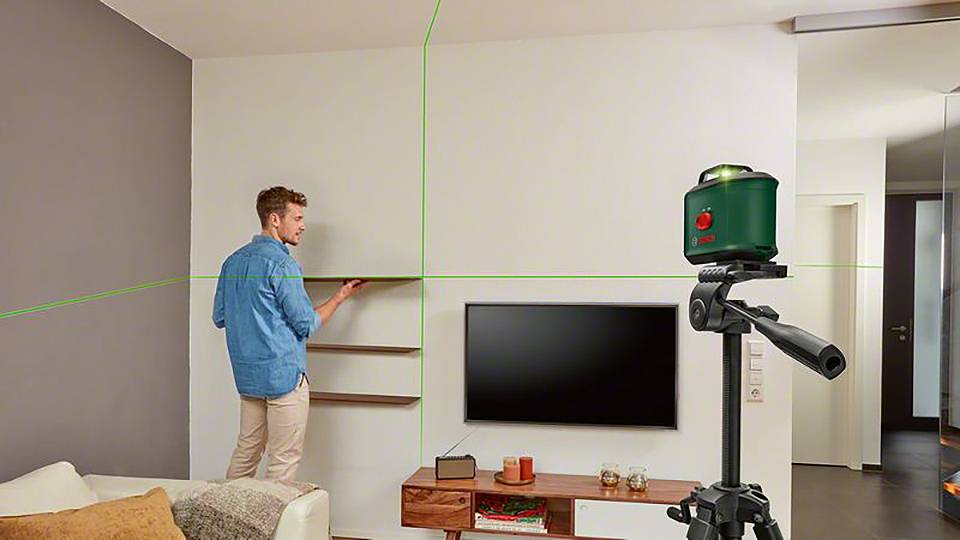 Muž zaměřuje police zeleným laserovým nivelačním přístrojem v obývacím pokoji. V pozadí je televize a pohovka.