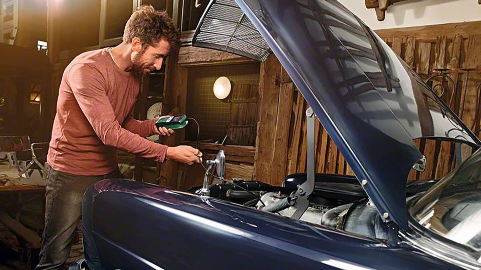 Muž kontroluje motor automobilu pomocí měřicího přístroje v autoservisu. Kapota je otevřená a vozidlo stojí v klidu.