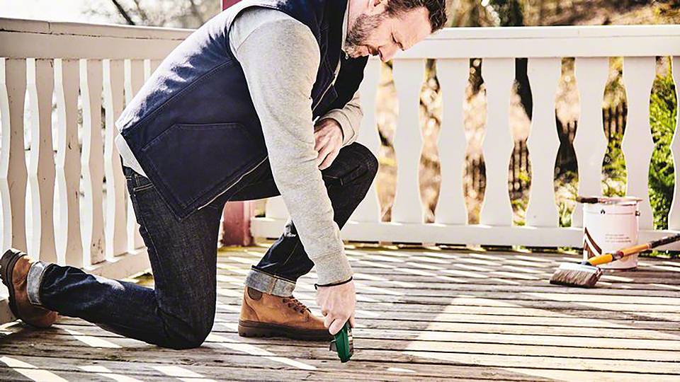 Muž klečí na dřevěné verandě a používá nástroj k úpravě podlahy. V pozadí stojí kbelík s barvou.
