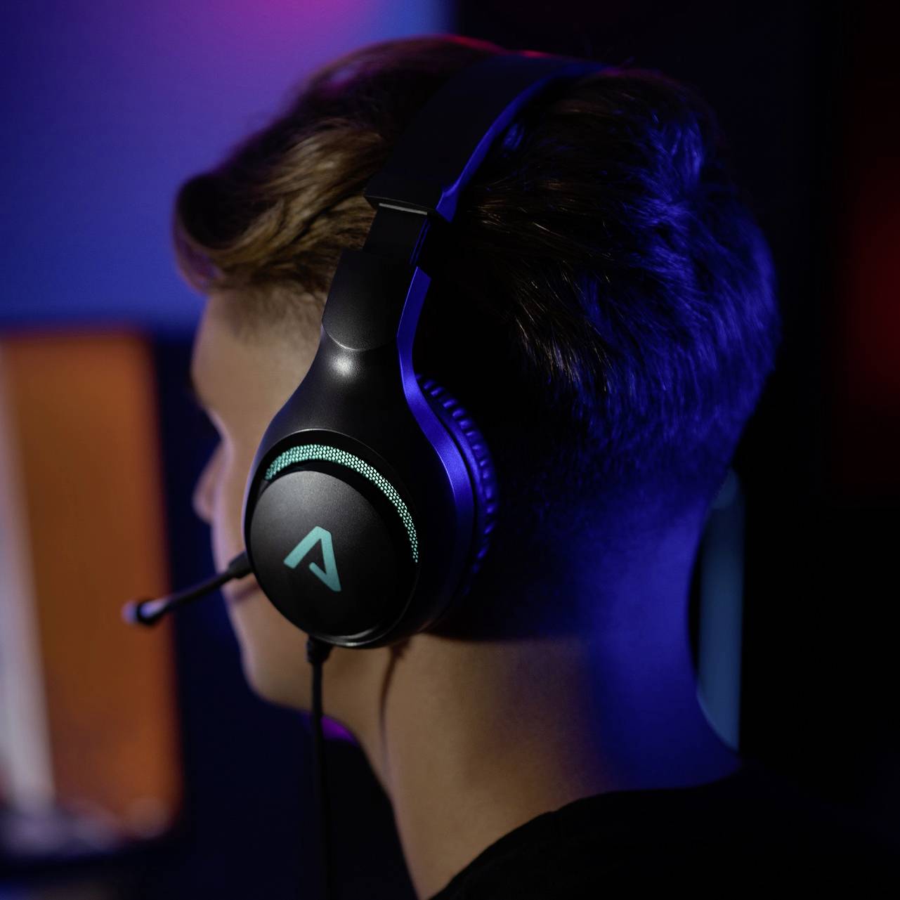 Osoba sedí v tlumeně osvětlené místnosti a nosí herní headset s mikrofonem. Logo na sluchátkách svítí modře.