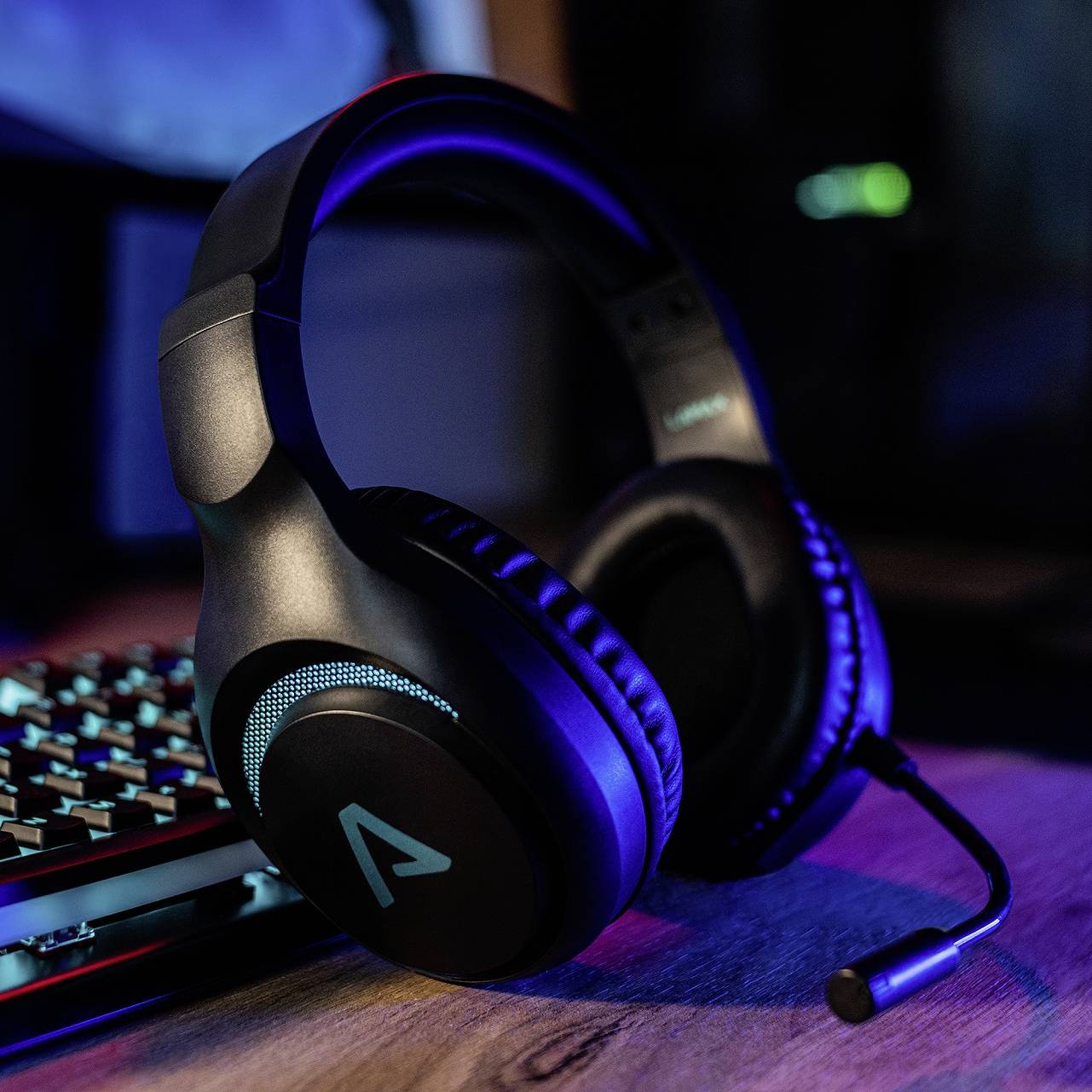 Herní headset leží na psacím stole vedle podsvícené klávesnice. Scéna je osvětlena modrým světlem.