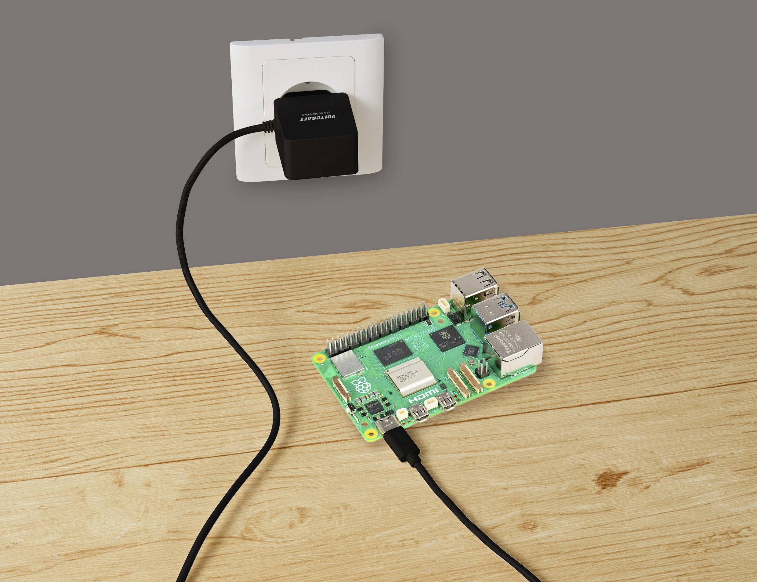 Počítačová deska Raspberry Pi na dřevěném stole, připojená k napájecímu adaptéru v nástěnné zásuvce.