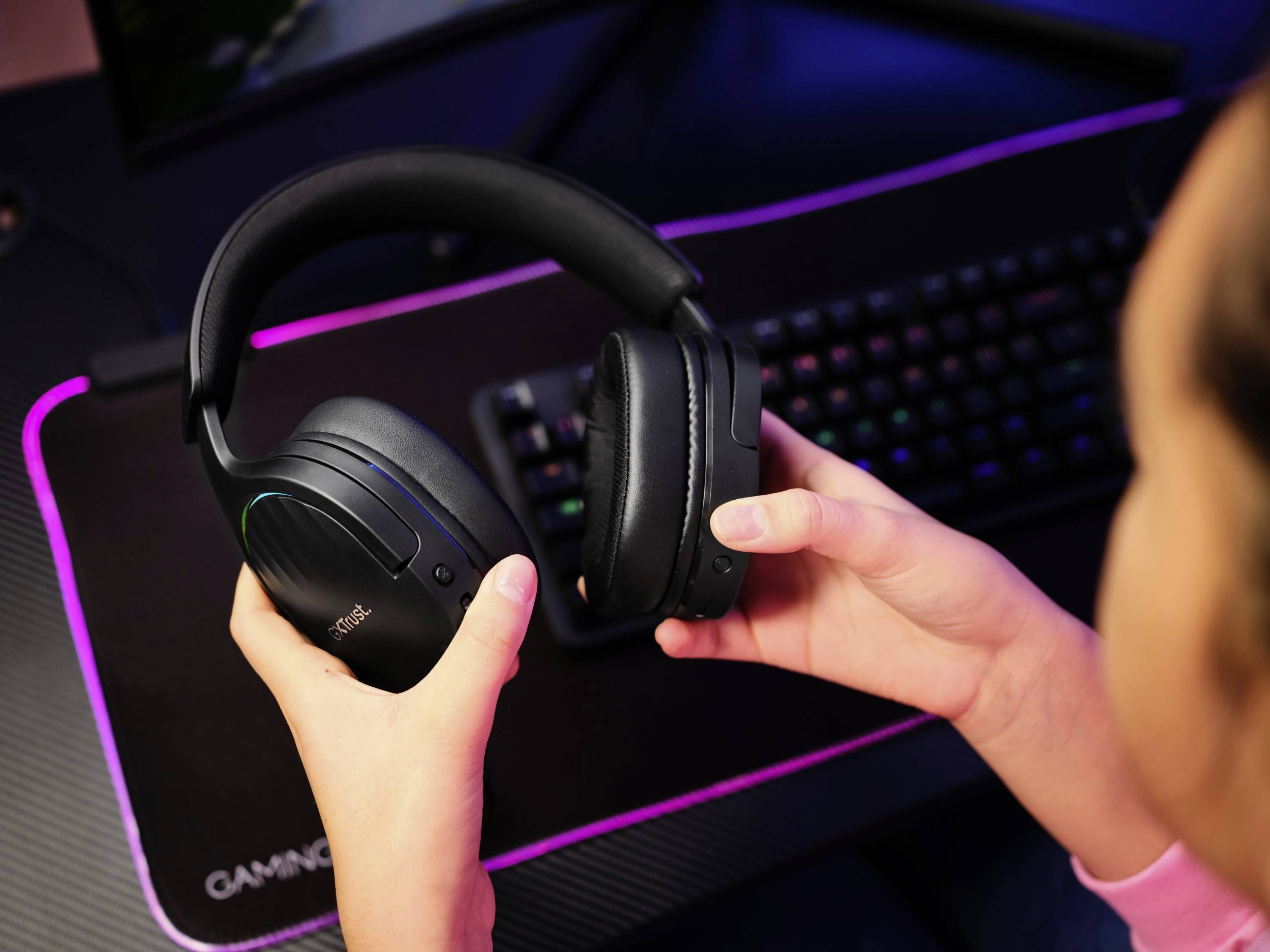 Osoba drží černou herní headset před osvětleným stolním počítačem s klávesnicí. Scéna naznačuje herní situaci.