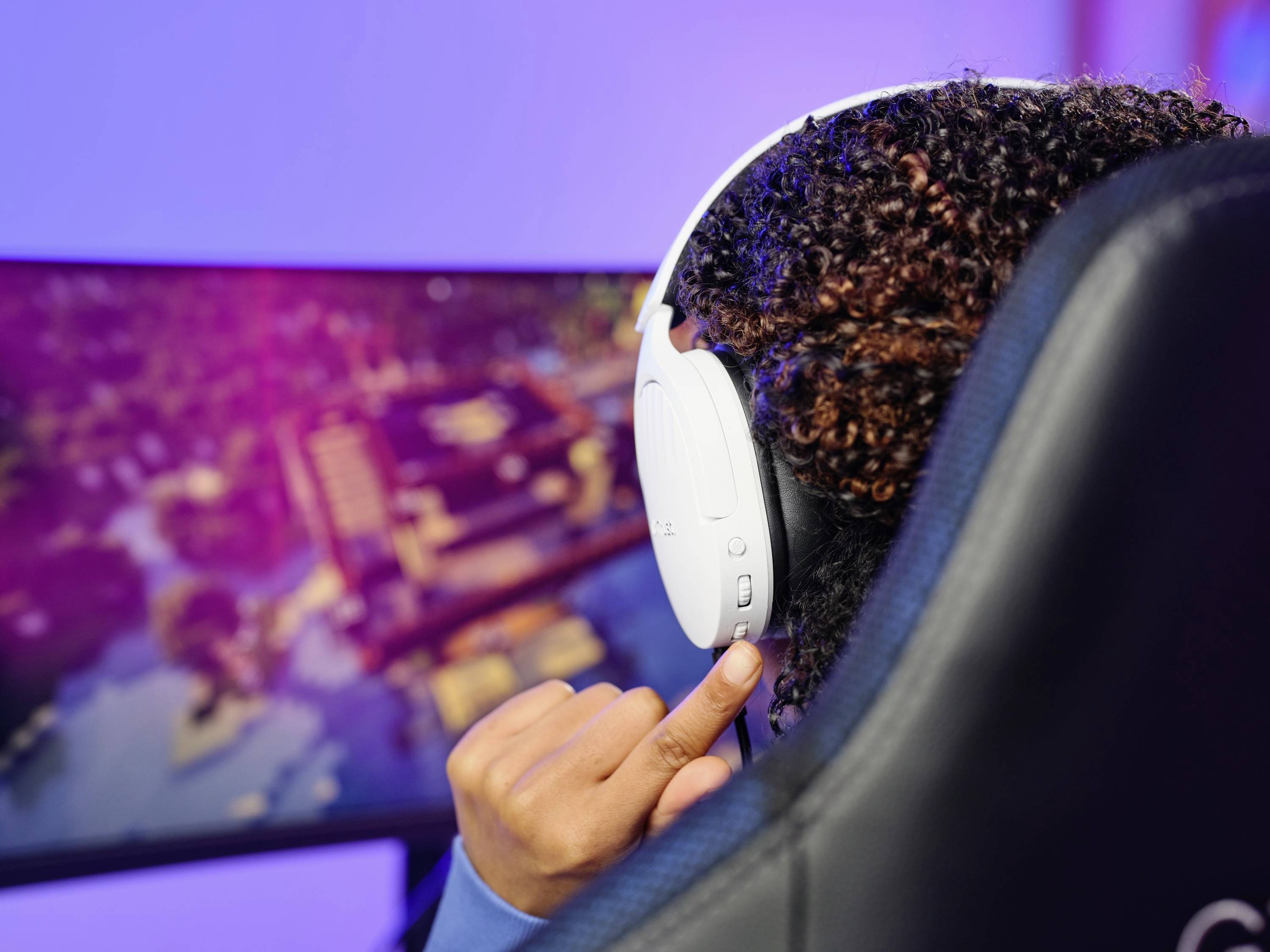 Osoba s bílými sluchátky hraje videohru na plochém monitoru. Obraz je zaměřen na herní zážitek.