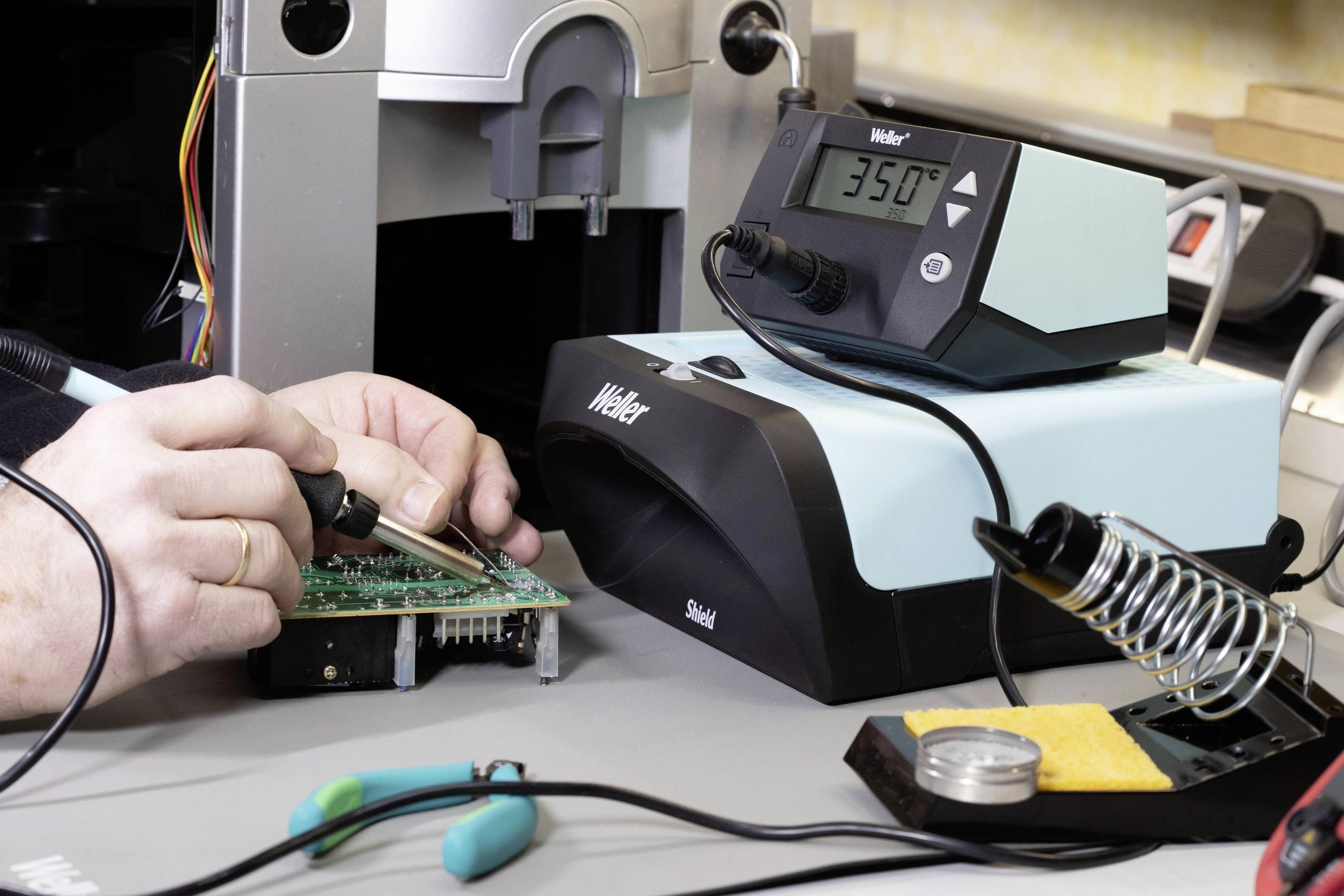Osoba pájí na desce plošných spojů pájecí pistolí, obklopena pájecími stanicemi a nástroji. Na obrazovce je zobrazeno '350°C'.