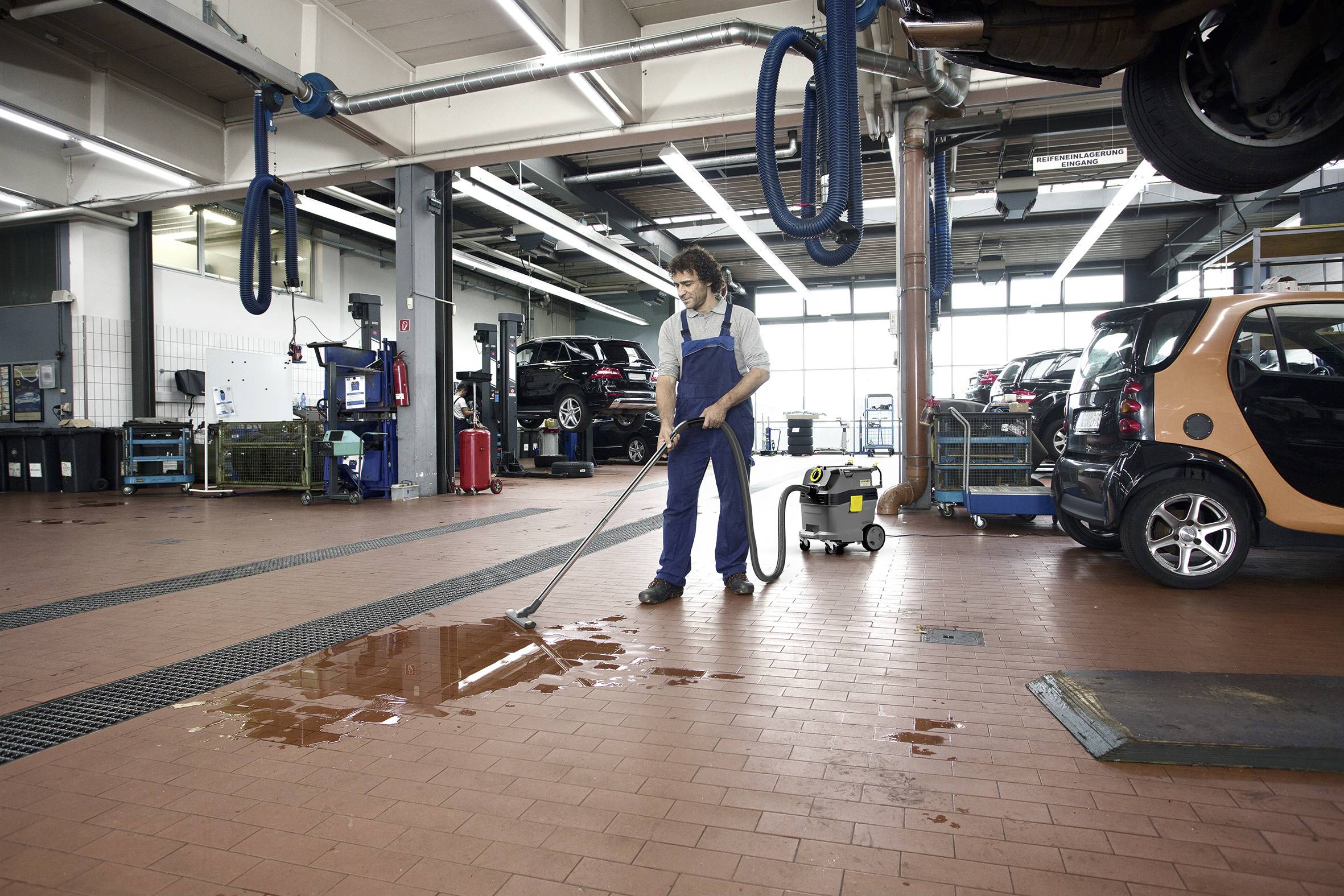 Muž v pracovním oblečení vysává louži na podlaze autobservisu. V pozadí stojí automobily.