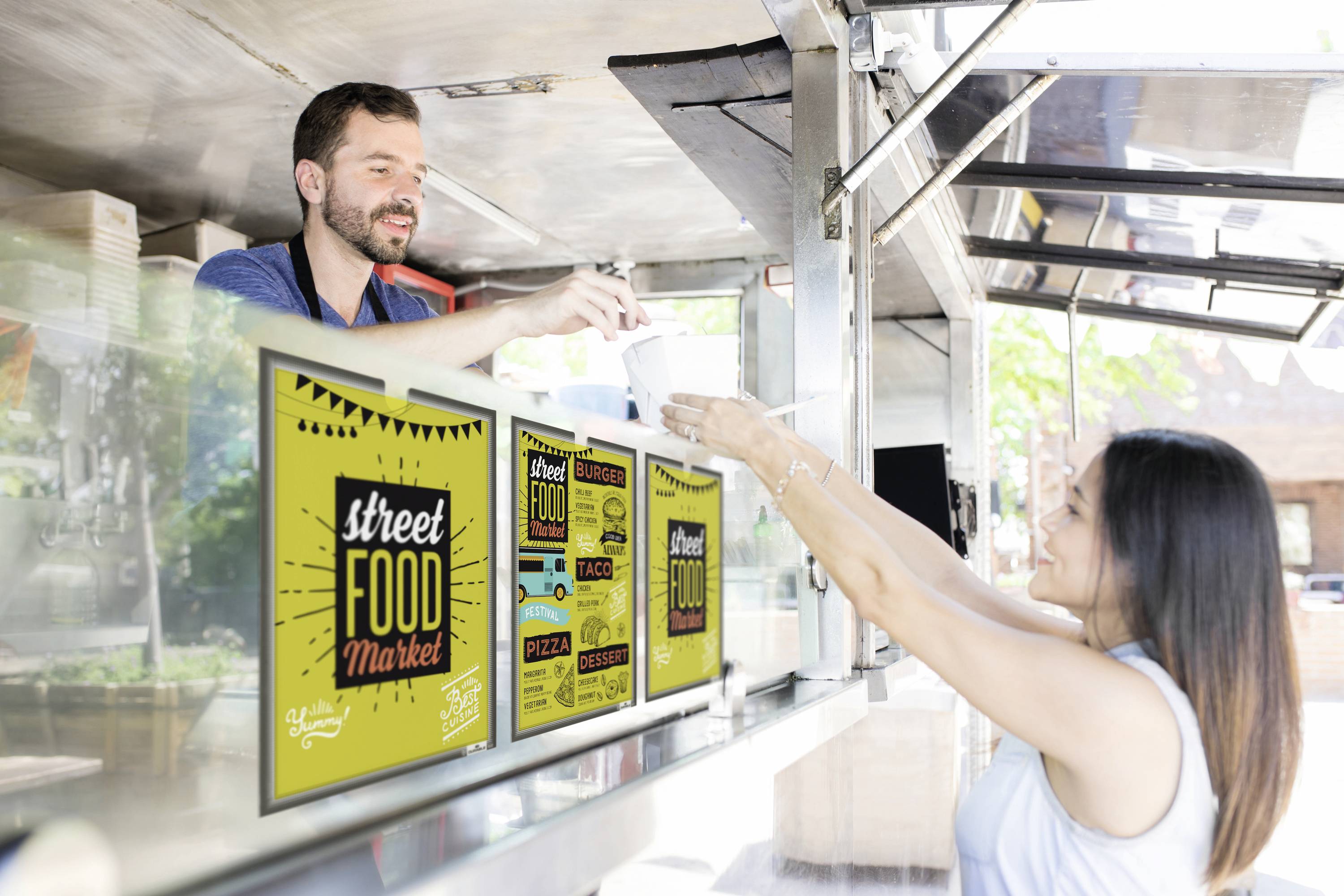 Muž v pojízdném stánku s občerstvením podává jídlo ženě. Cedule na voze propagují 'Street Food Market' s různými pokrmy.