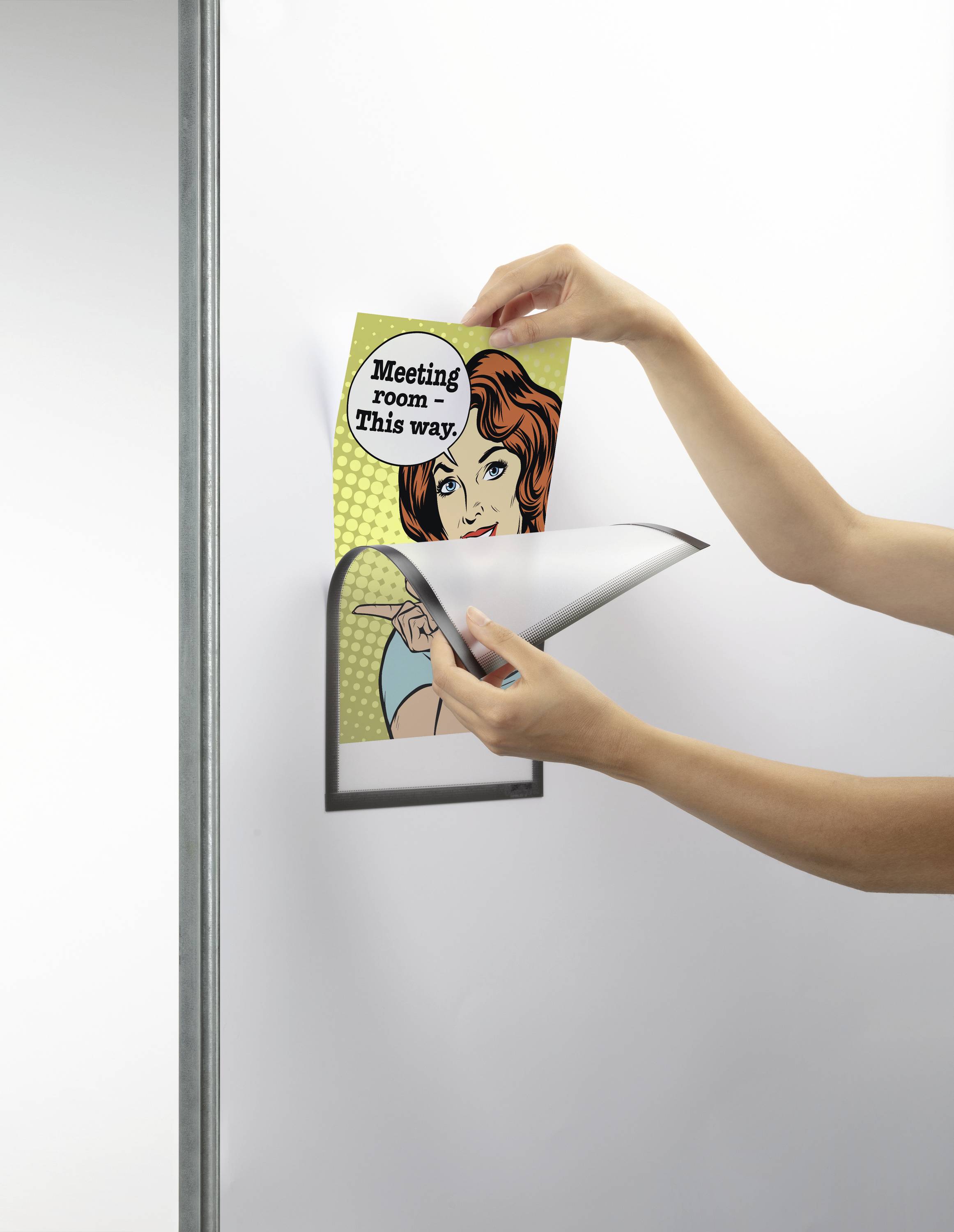 Osoba mění plakát s nápisem 'Meeting room this way' v nástěnném rámu. Plakát zobrazuje komiksovou postavu.