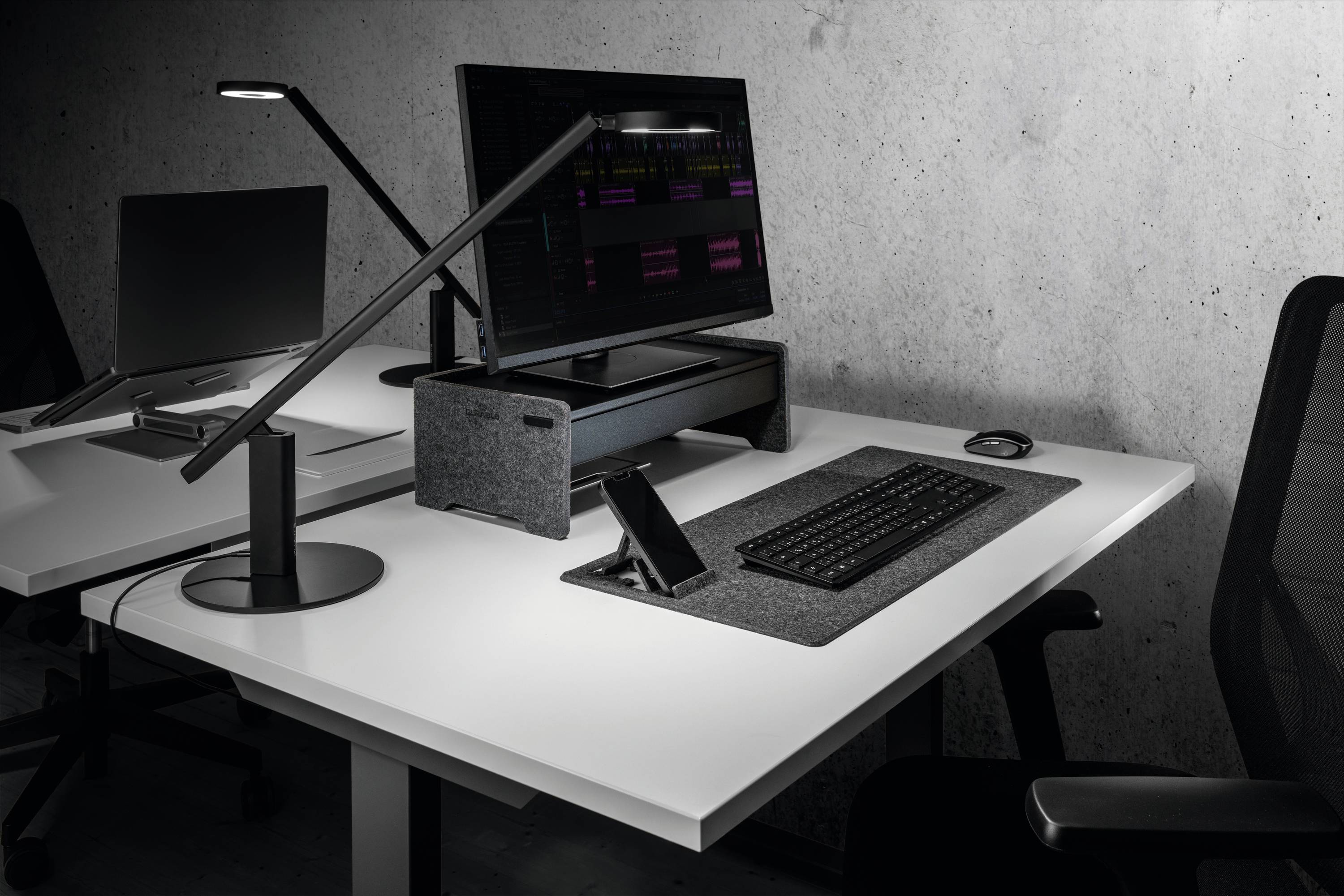 Moderní psací stůl s počítačem, klávesnicí a lampou. Stůl je uspořádaný a navržený v minimalistickém stylu.