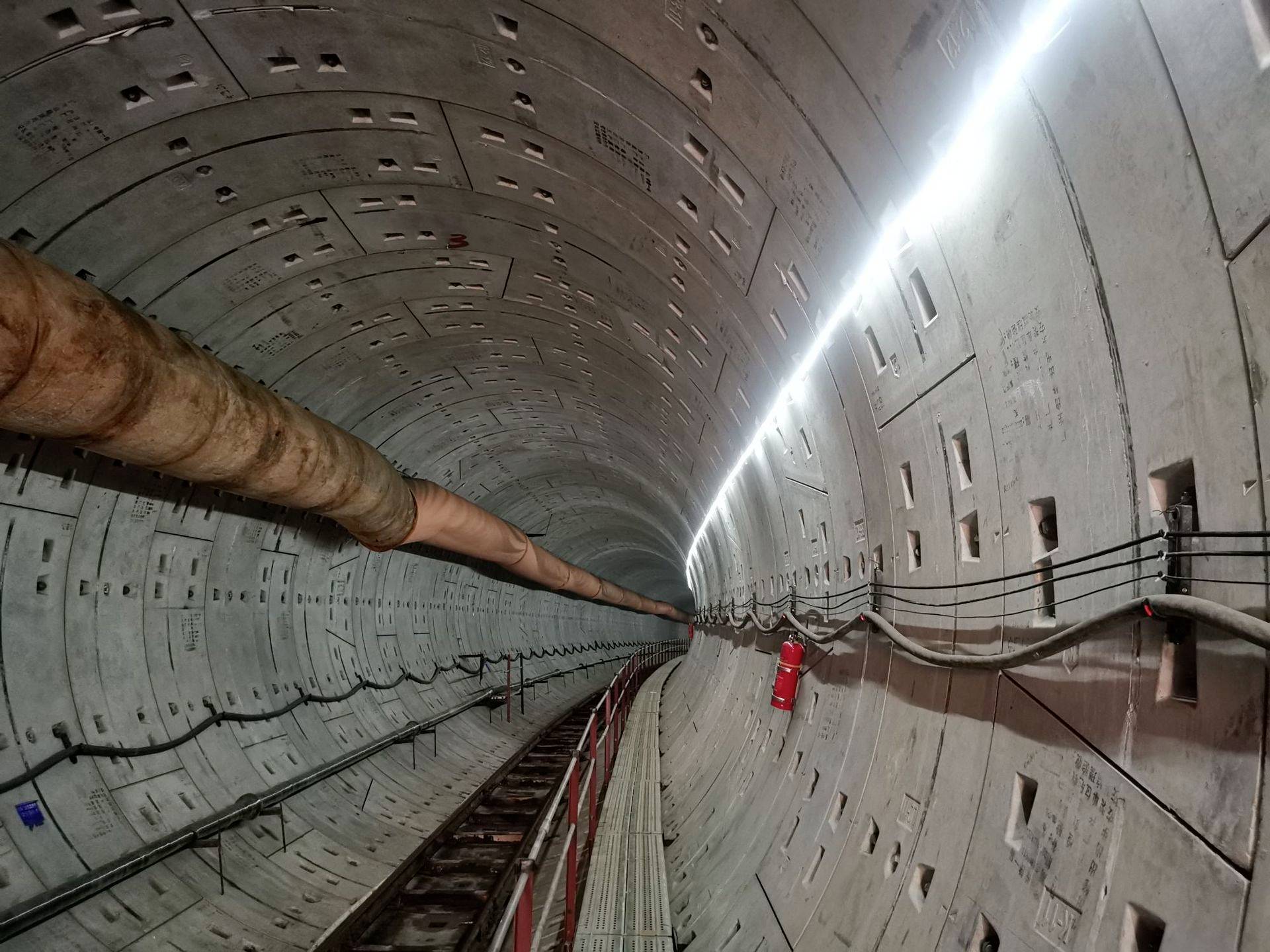 Pohled do betonového metrového tunelu s osvětlením, trubkami a kabely na stěnách. Je zde viditelný hasicí přístroj.