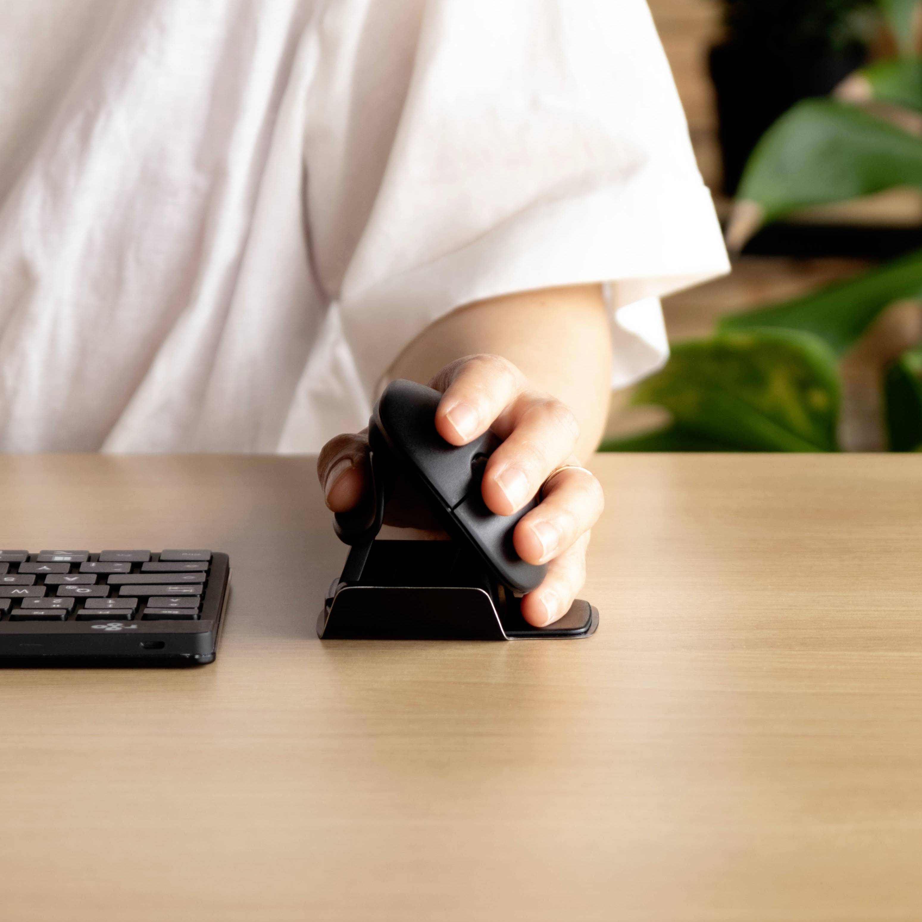 Osoba píše na ergonomické klávesnici a používá vertikální myš. V pozadí jsou viditelné rostliny.