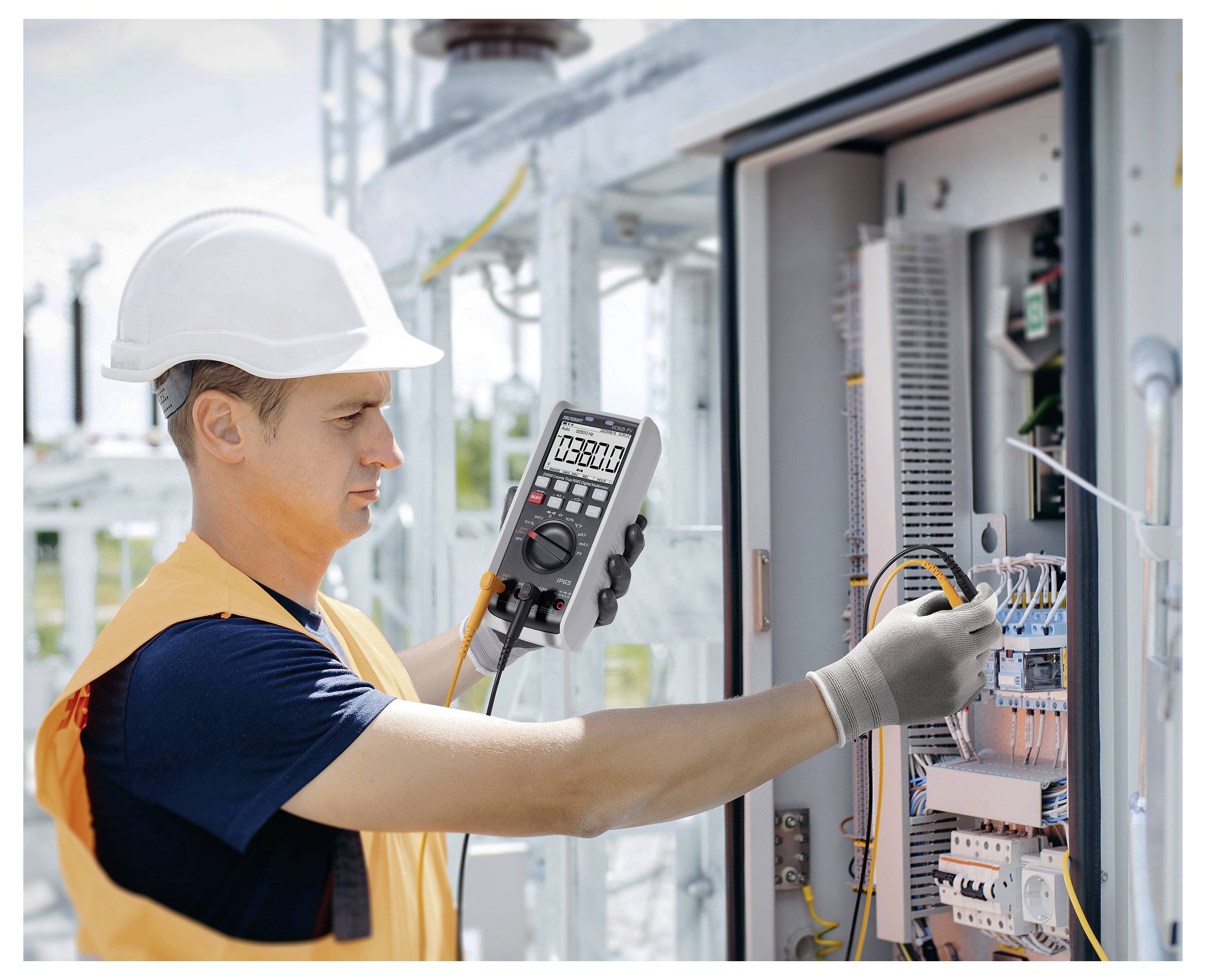 Elektrikář v ochranné přilbě testující elektroinstalaci v otevřeném elektrickém rozvaděči digitálním multimetrem.