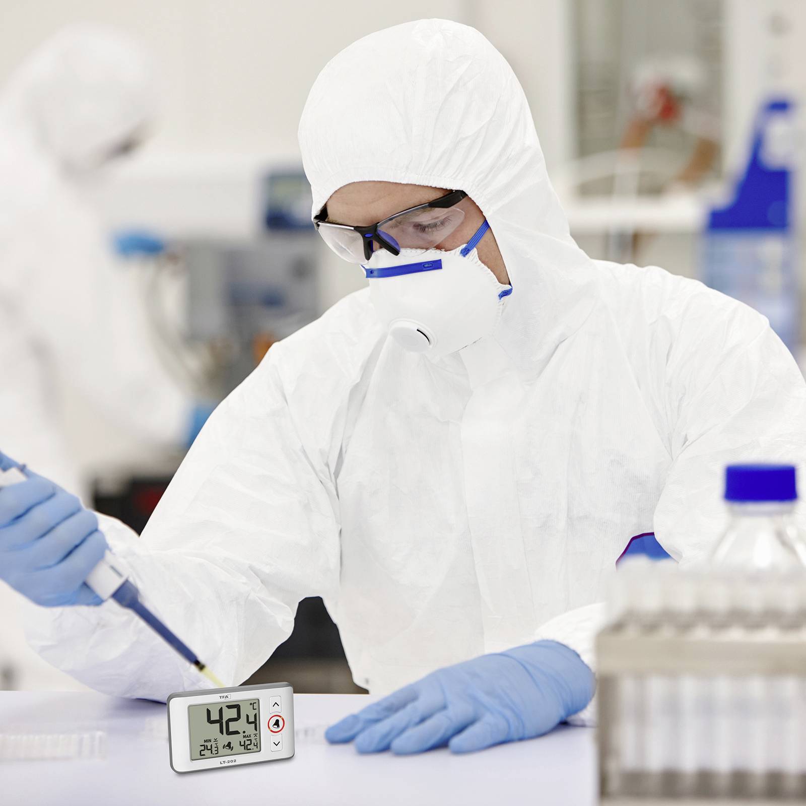 Osoba v ochranném obleku pracuje v laboratoři a pipetuje tekutinu. Digitální teploměr ukazuje 42,4 °C. Vědecké výzkumné prostředí.