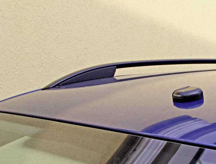 Modré auto s příčníky na střeše a malou anténou na střeše před krémovou zdí.