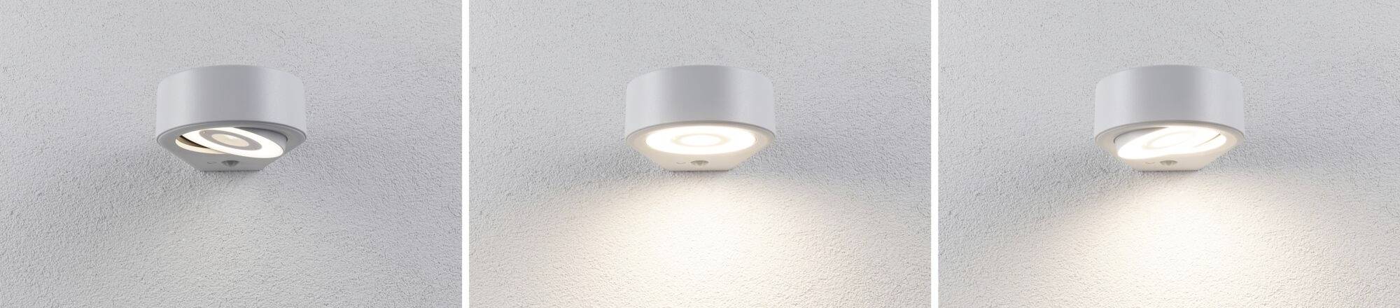 Tři bílé nástěnné lampy na šedé stěně. Každá lampa září s jinou intenzitou světla: vpravo nejjasnější, vlevo nejslabší.