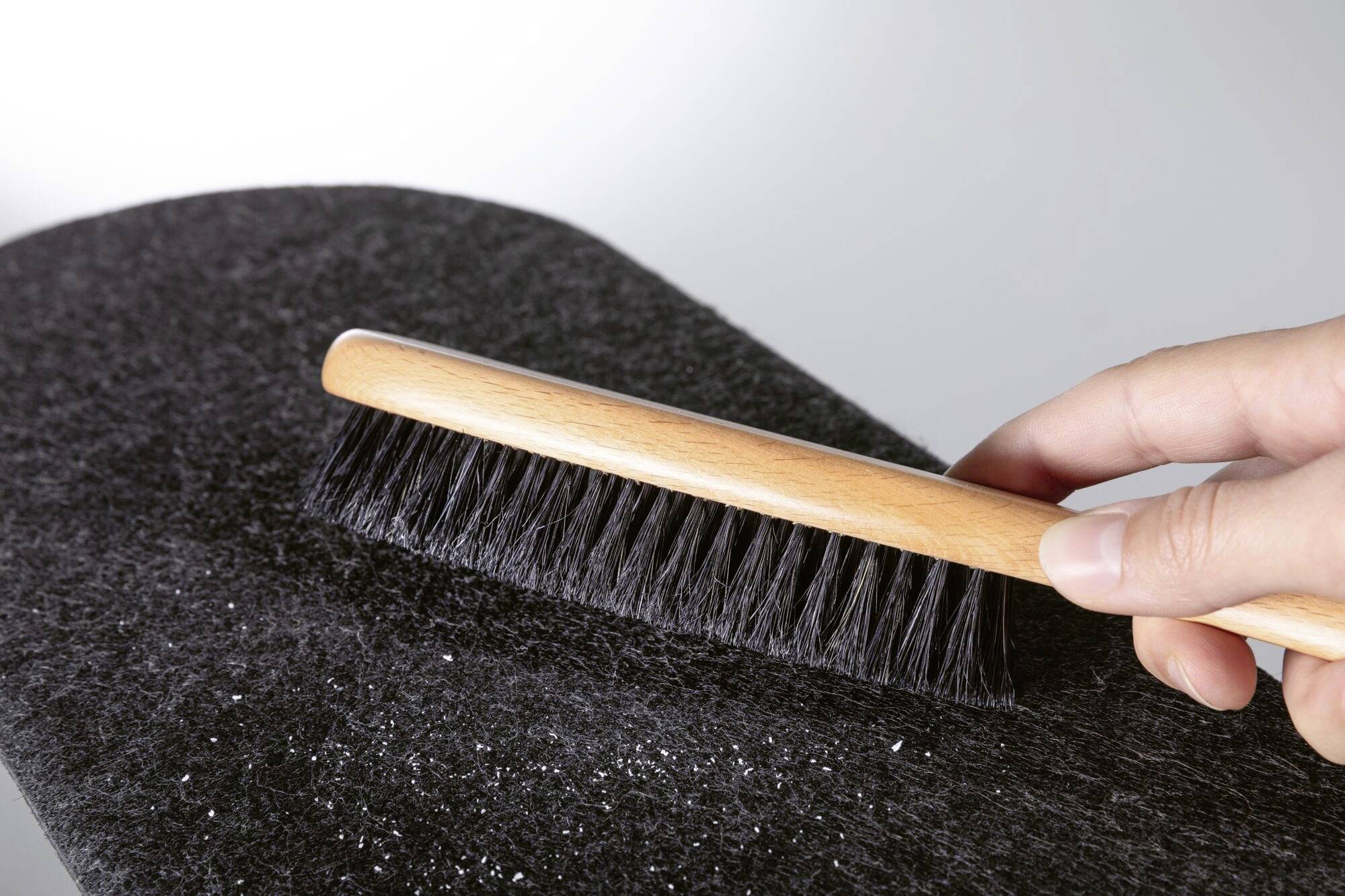 Jedna ruka drží štětku s dřevěnou rukojetí a černými štětinami, čistí tmavou látku nebo koberec, odstraňuje viditelné drobky.