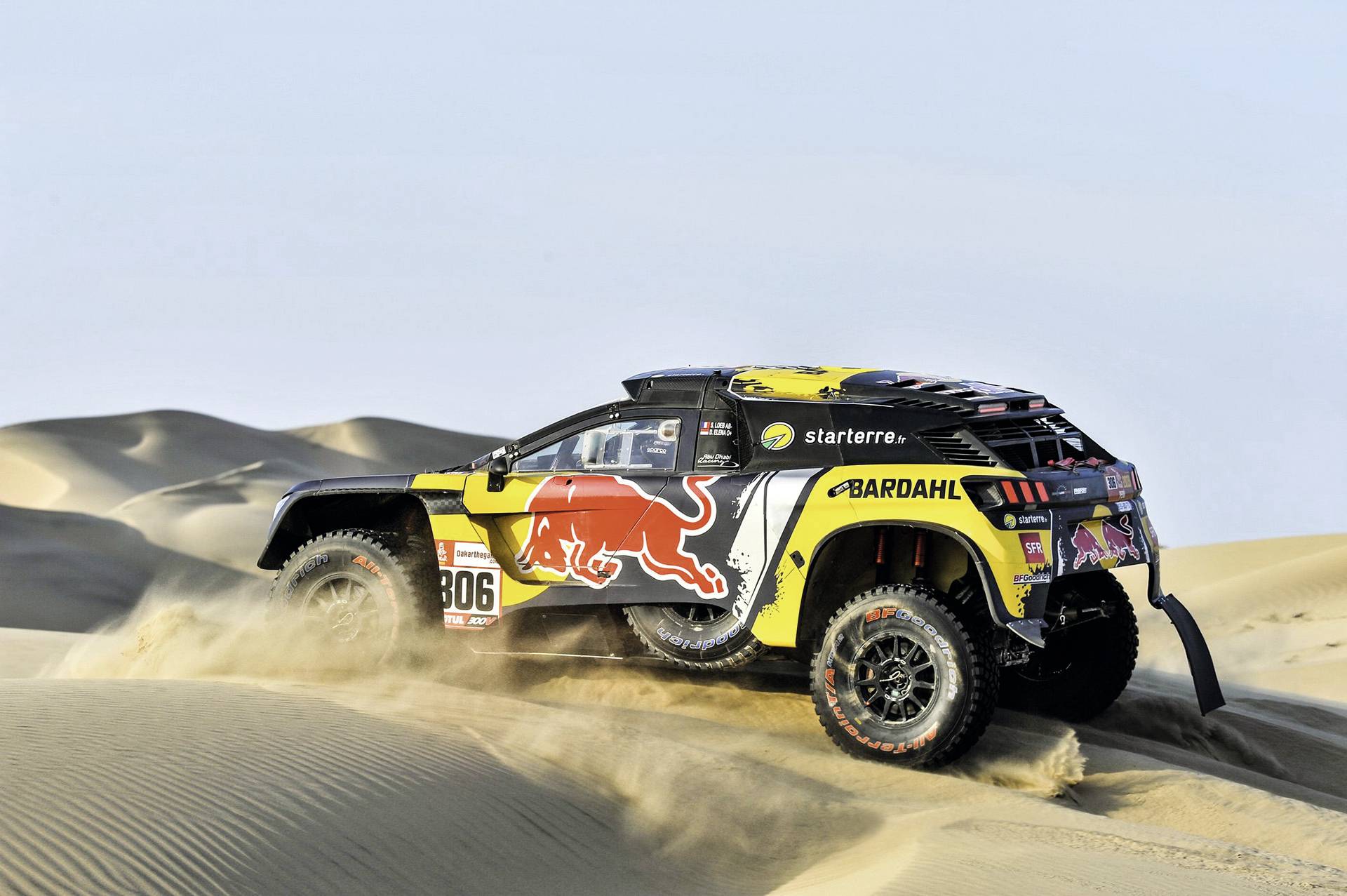 Žluté rally auto s logy 'Bardahl' a 'Red Bull' projíždí pouštní krajinou s vysokými písečnými dunami.