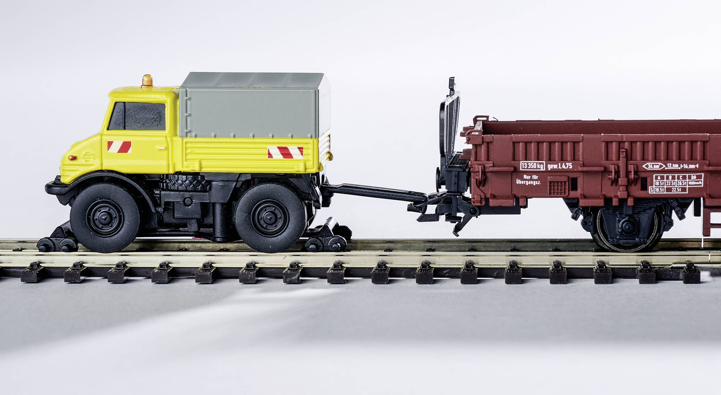 Žlutý modelový náklaďák táhnehnědý přívěs po kolejích modelové železnice. Přívěs má nápisy.