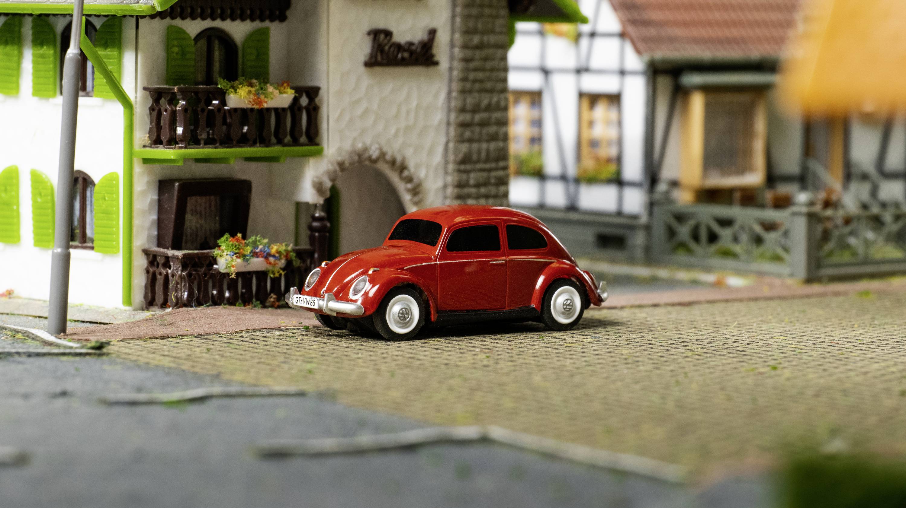 Červené hračkářské auto je zaparkované před miniaturním hrázděným domem s zelenými balkóny a truhlíky s květinami. Scéna v modelovém měřítku.