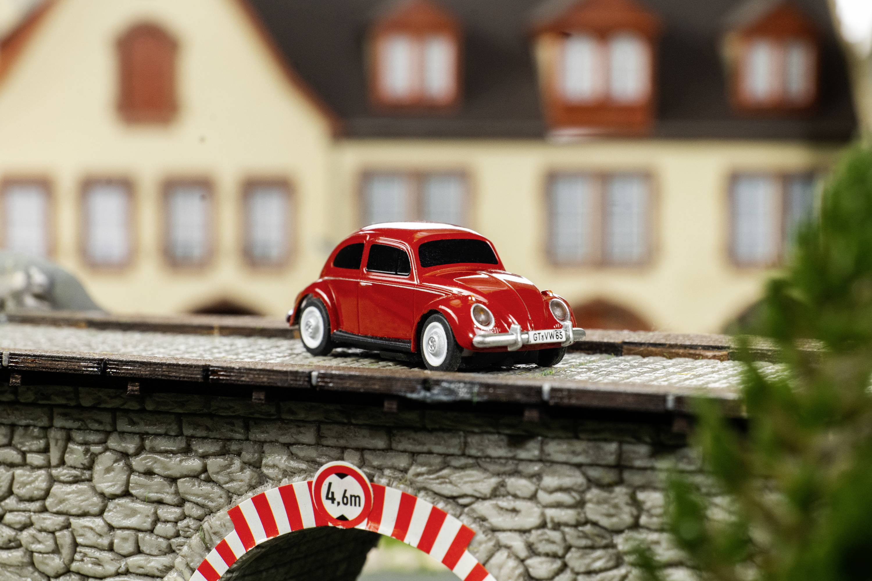 Červené hračkářské autíčko na miniaturním kamenném mostě před modelovým domem. Cedule ukazuje '4,6 m'.