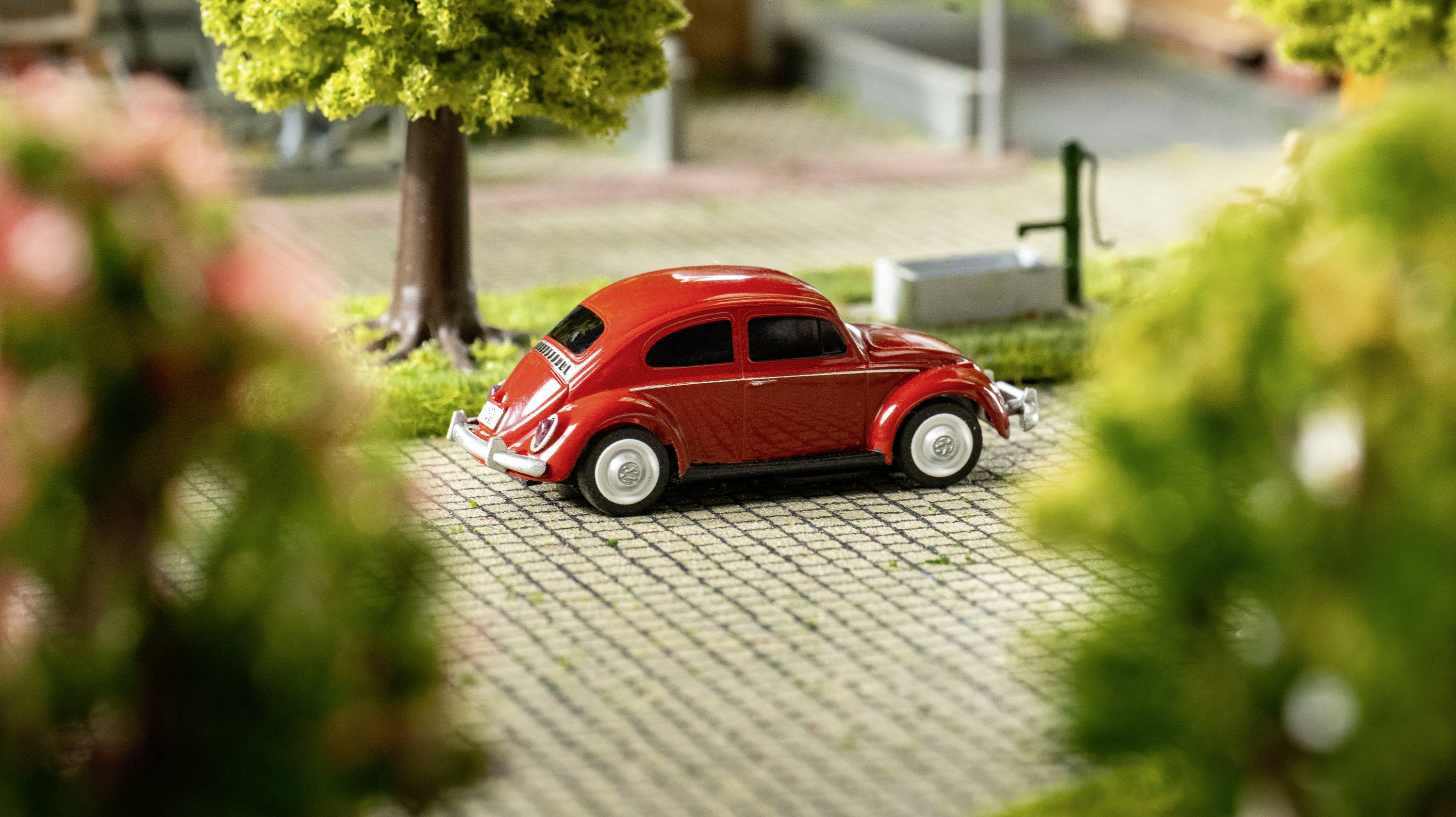 Červené hračkářské auto ve stylu veterána stojí na dlážděné silnici, obklopené stromy a rozmazaným pozadím.