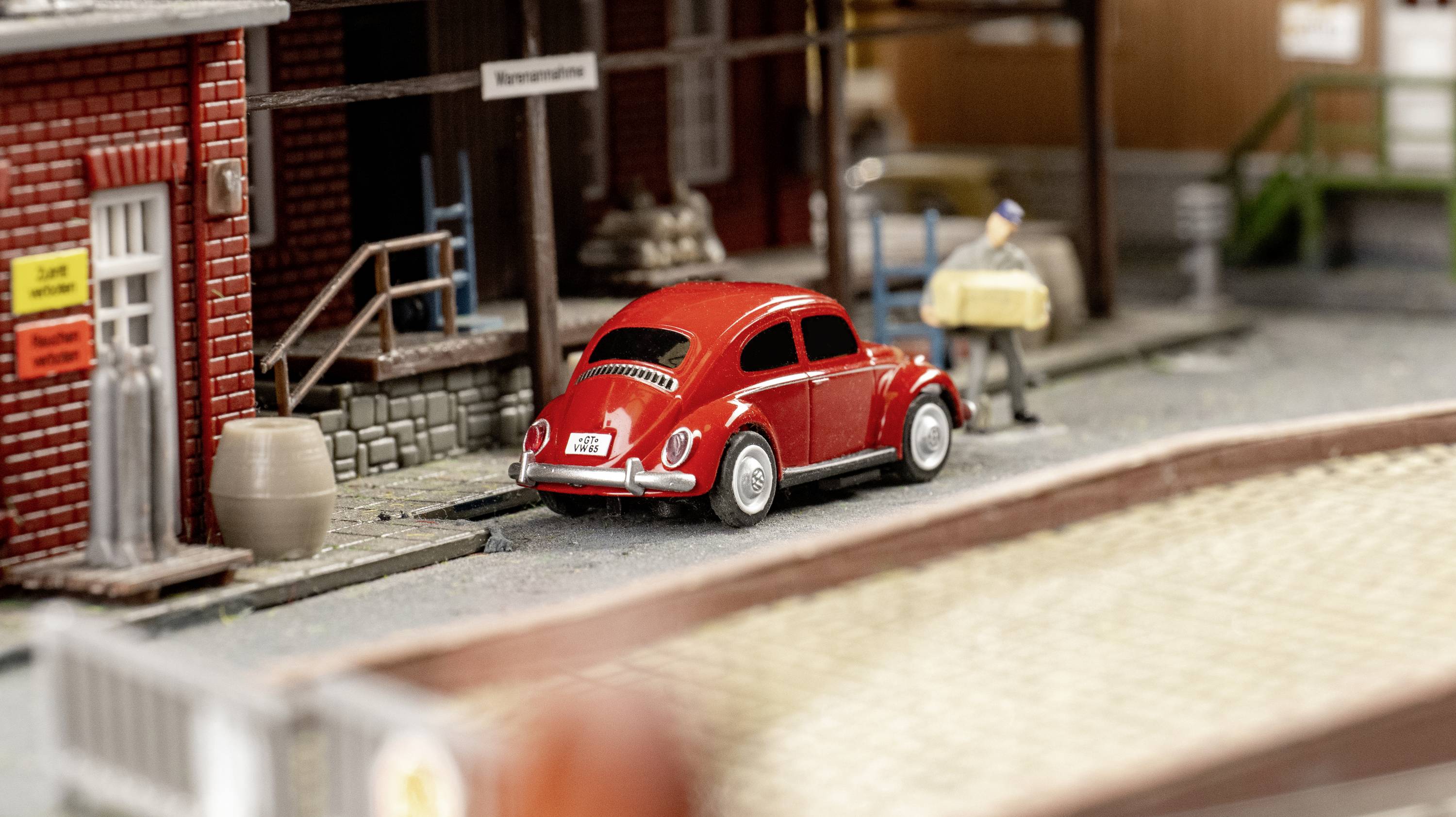 Červený VW Brouk je zaparkovaný vedle cihlové budovy na miniaturní silnici. Vedle něj stojí miniaturní člověk.