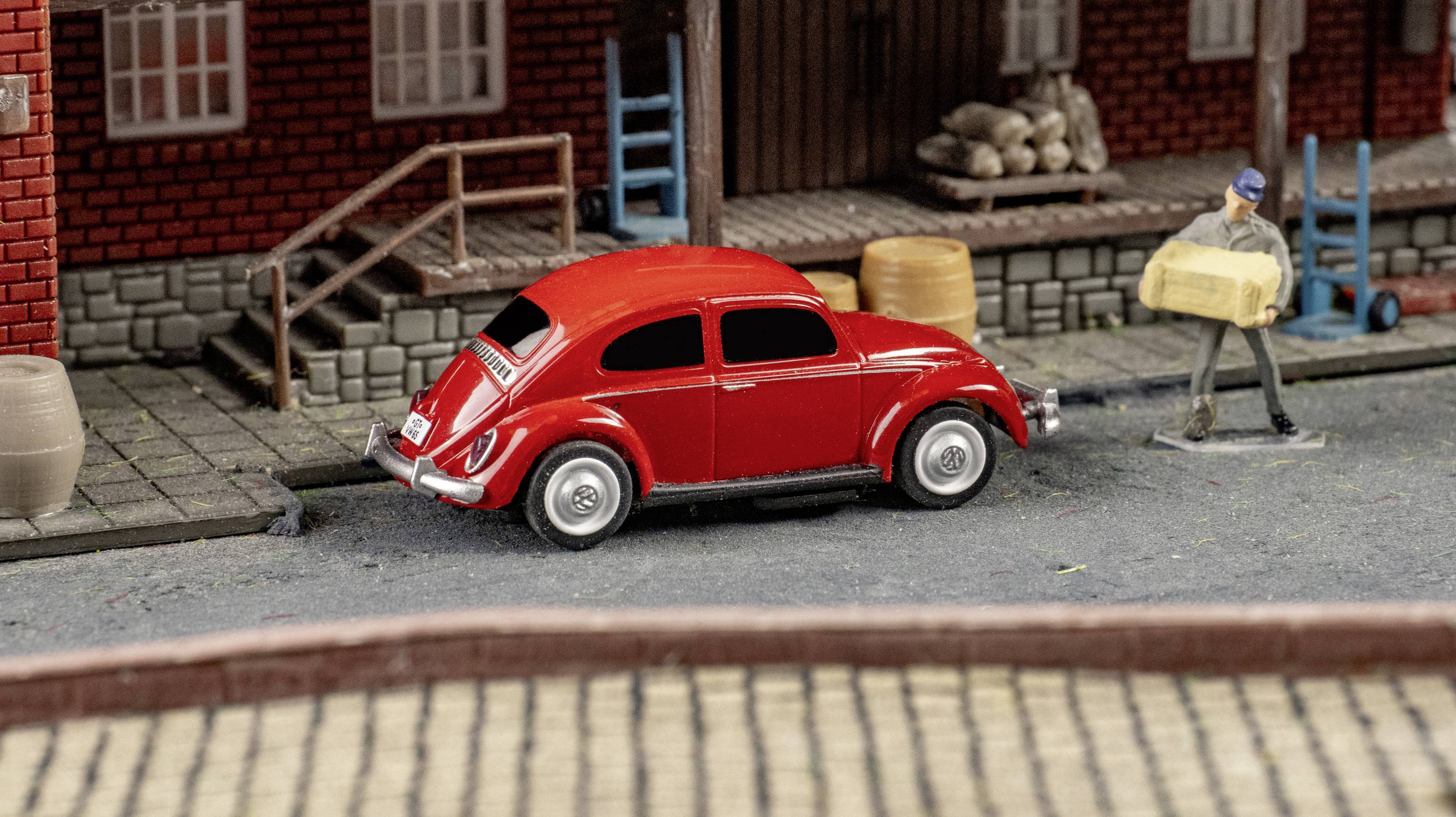 Červené hračkářské auto na modelové silnici před cihelnou budovou, vedle stojí postava nesoucí velkou krabici.